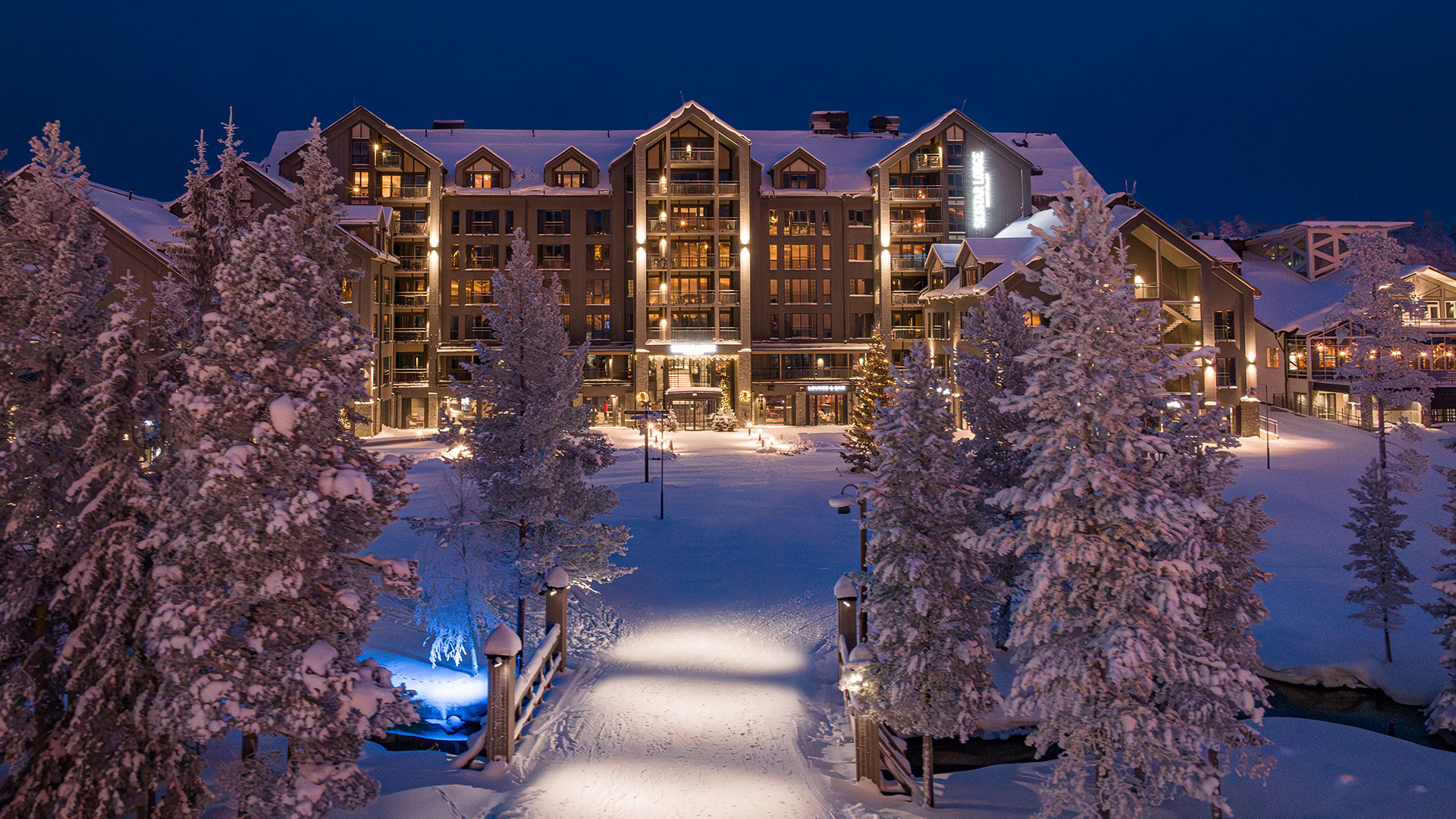 Hotell i vinterlandskap, upplyst på kvällen, omgivet av snötäckta träd