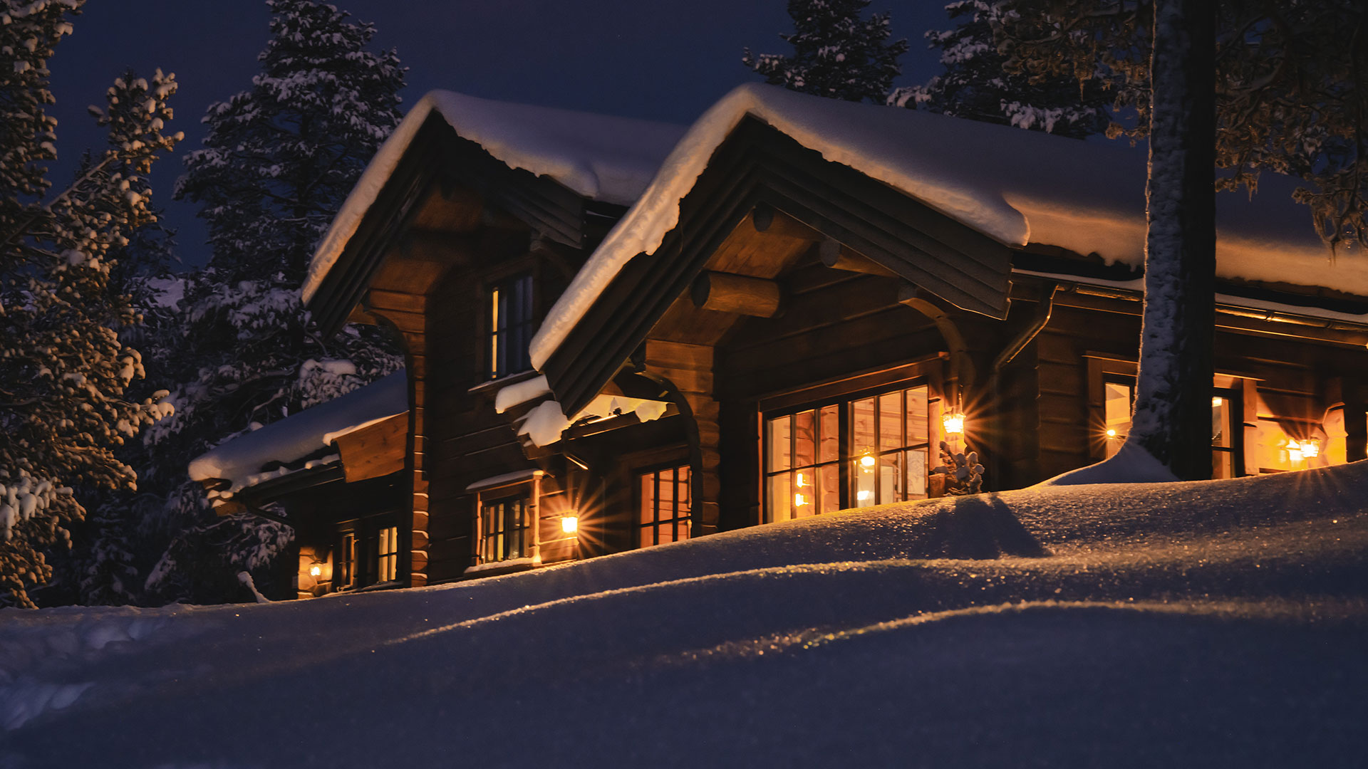 Traditionell timmerstuga täckt av snö och omgiven av snötäckta träd. Varmt mysigt lyser upp stugan inifrån.