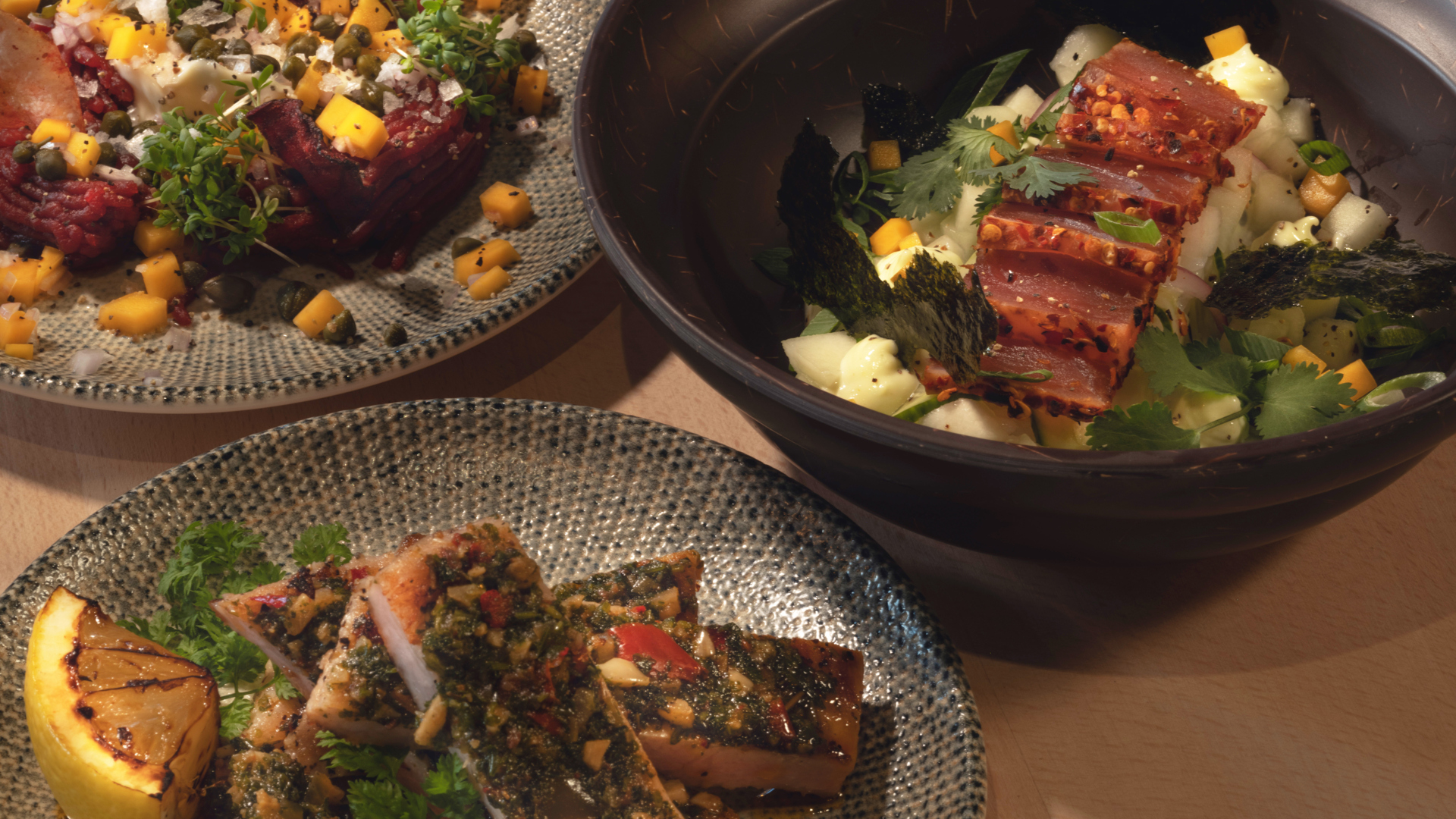 Tre tallrikar med färgglada, konstnärligt upplagda maträtter, inklusive krispiga grönsaker och kryddig tonfisk, serverade med färska örter och grillad citron