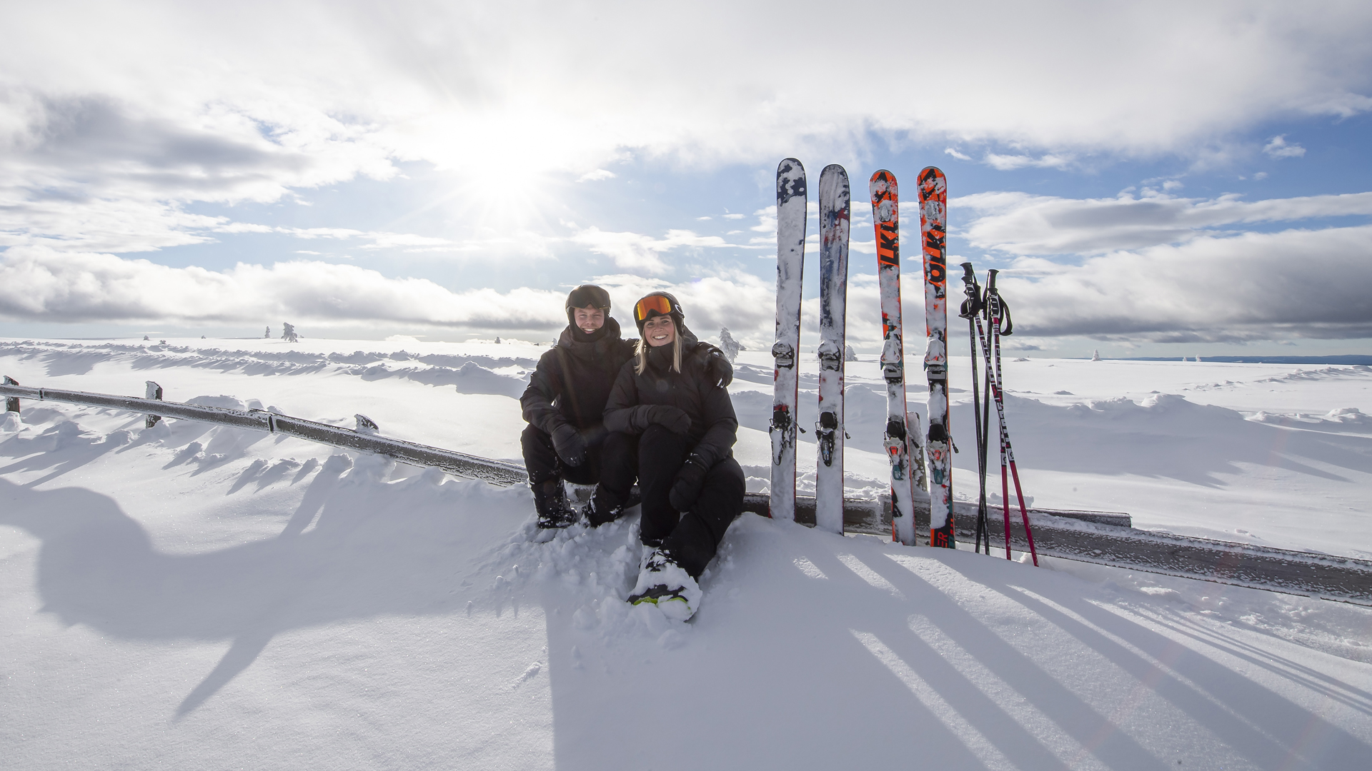 Två personer sitter i snön med skidor och stavar i ett vintrigt landskap, solig dag med moln på himlen
