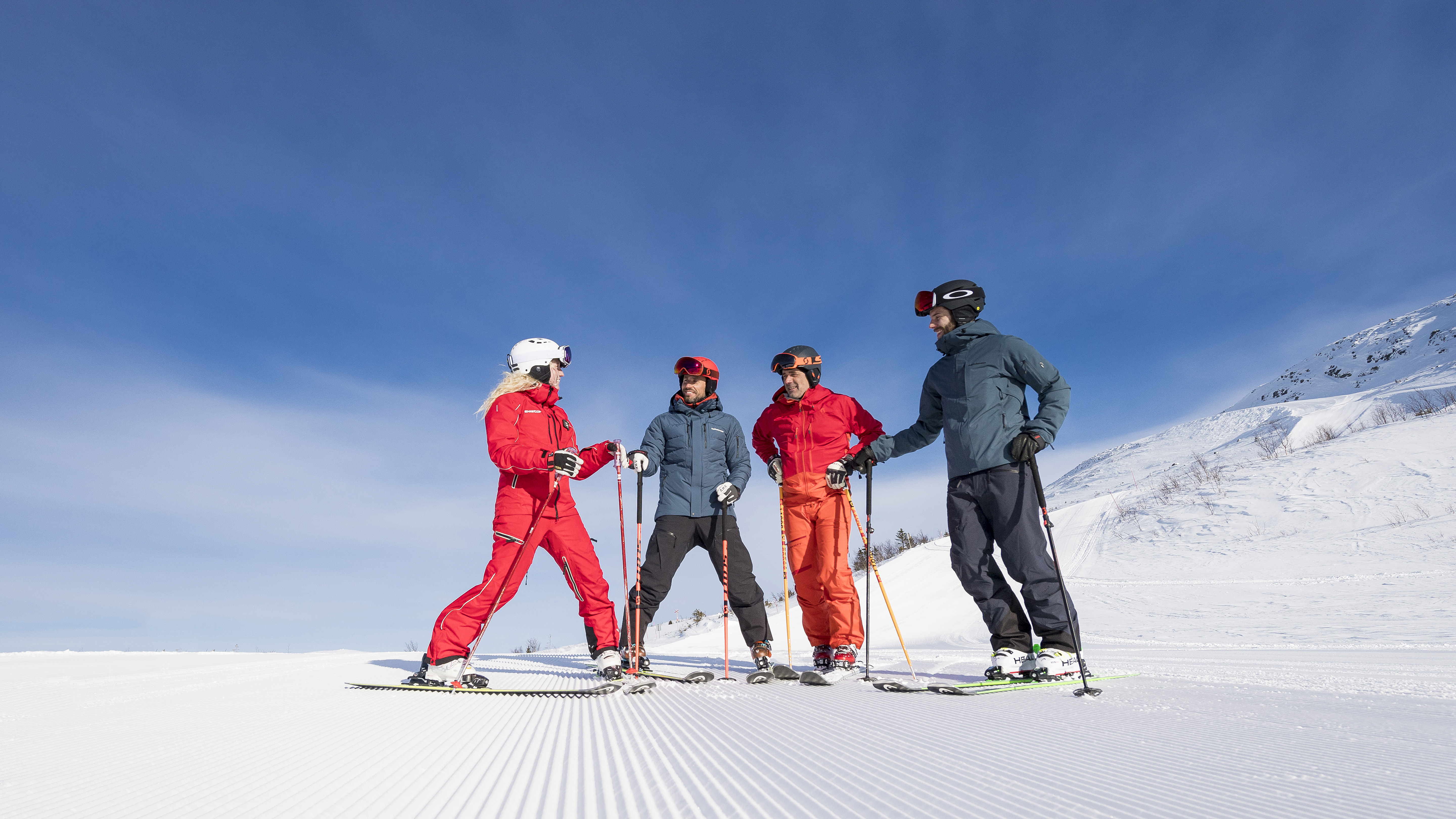Fyra personer klädda i skidkläder och hjälmar står tillsammans på en snötäckt skidbacke under en klarblå himmel