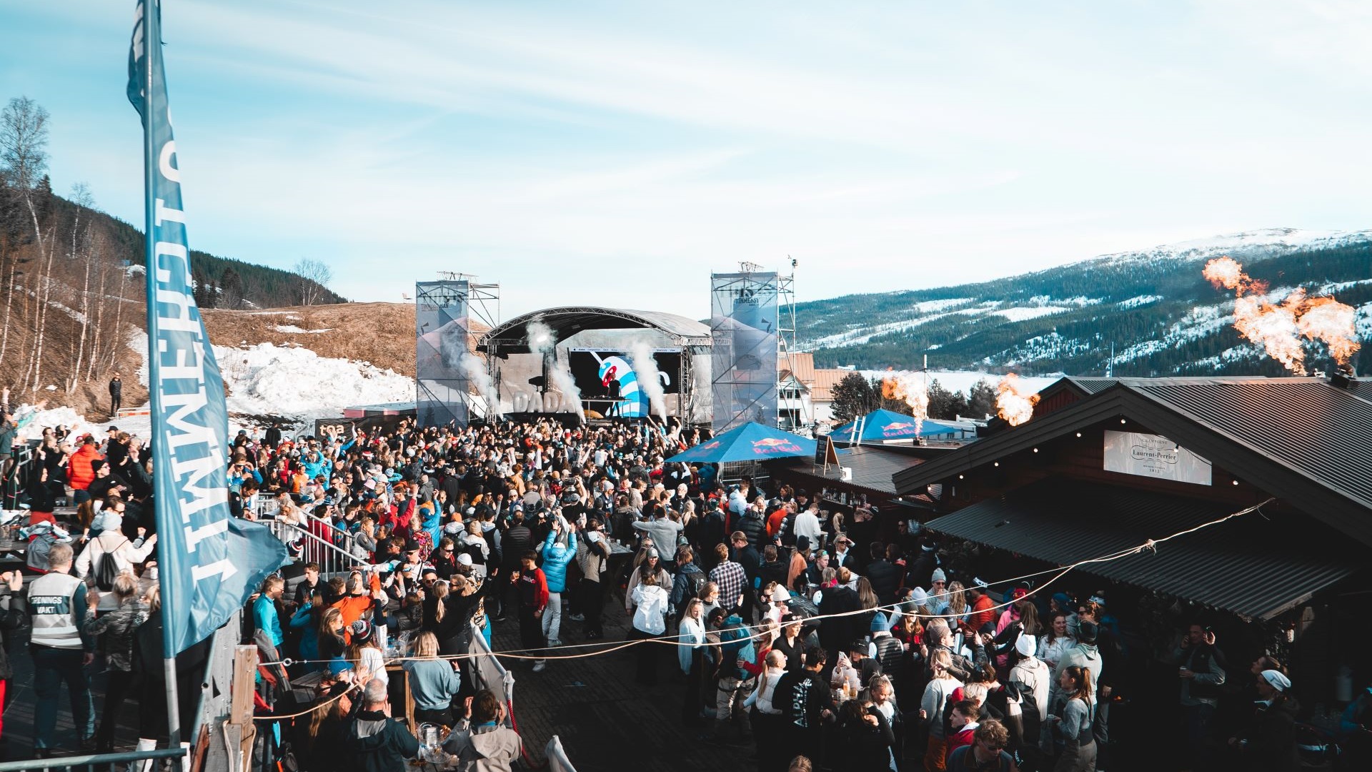 Utendørskonsert i skisted med stort publikum, snødekte fjell i bakgrunnen og scener med fargerike flagg