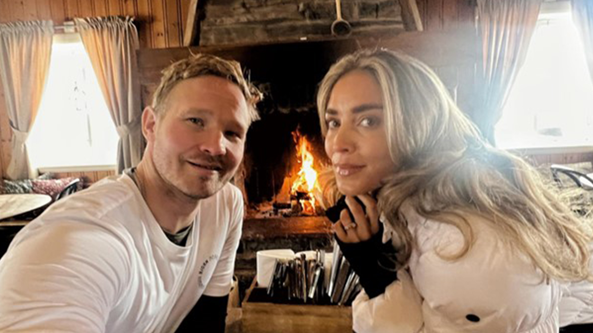 Man och kvinna framför en eldstad i en mysig stuga, klädda i vinterkläder