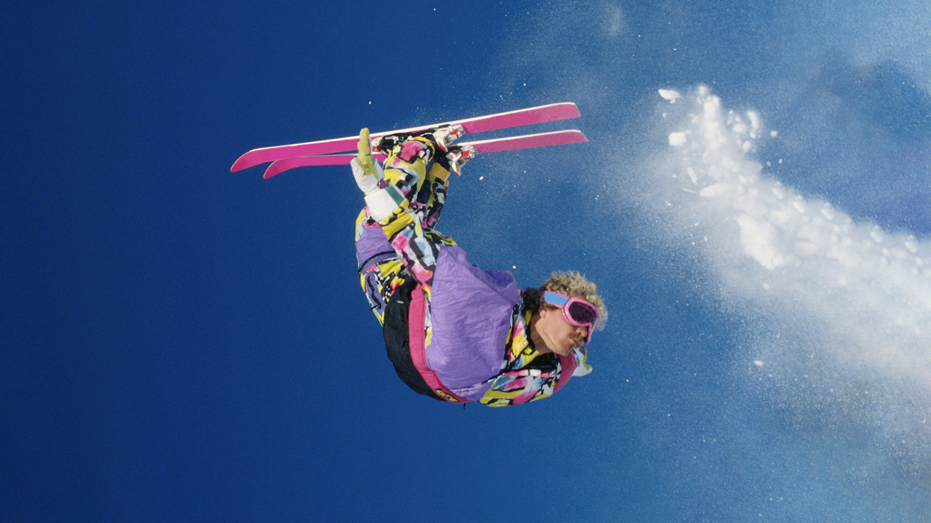 Skidåkare i färgglad dräkt utför ett trick med rosa skidor mot en klarblå himmel