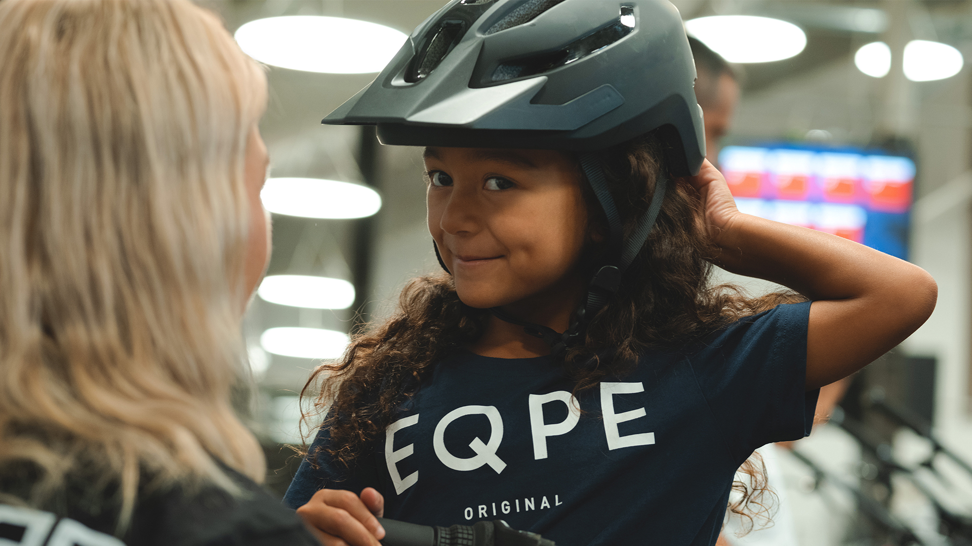 Flicka med mörkt lockigt hår justerar sin hjälm och ler mot en vuxen person i en ljusfylld inomhusmiljö. Hon bär en mörkblå tröja med texten "EQPE"
