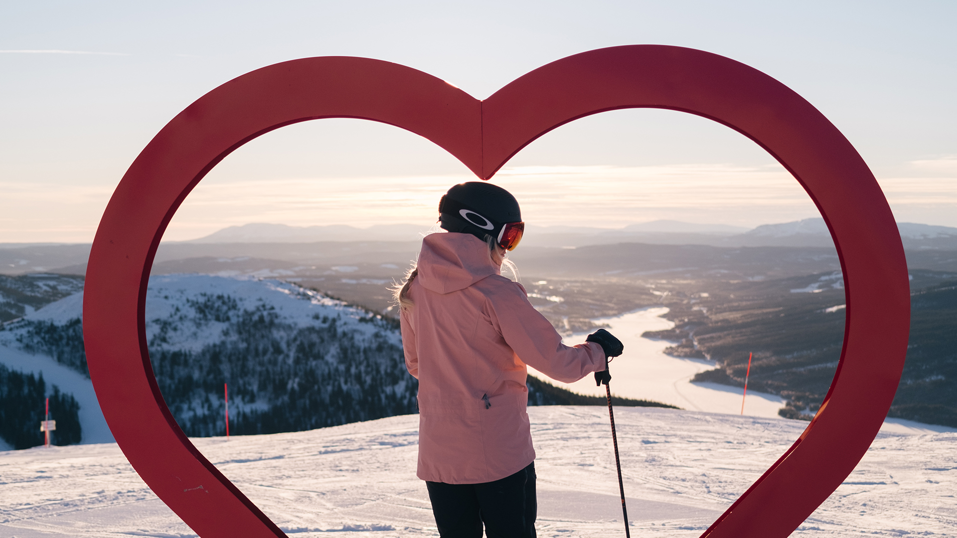 Person står på en snötäckt bergstopp och ser ut över ett vackert vinterlandskap genom en hjärtformad skulptur