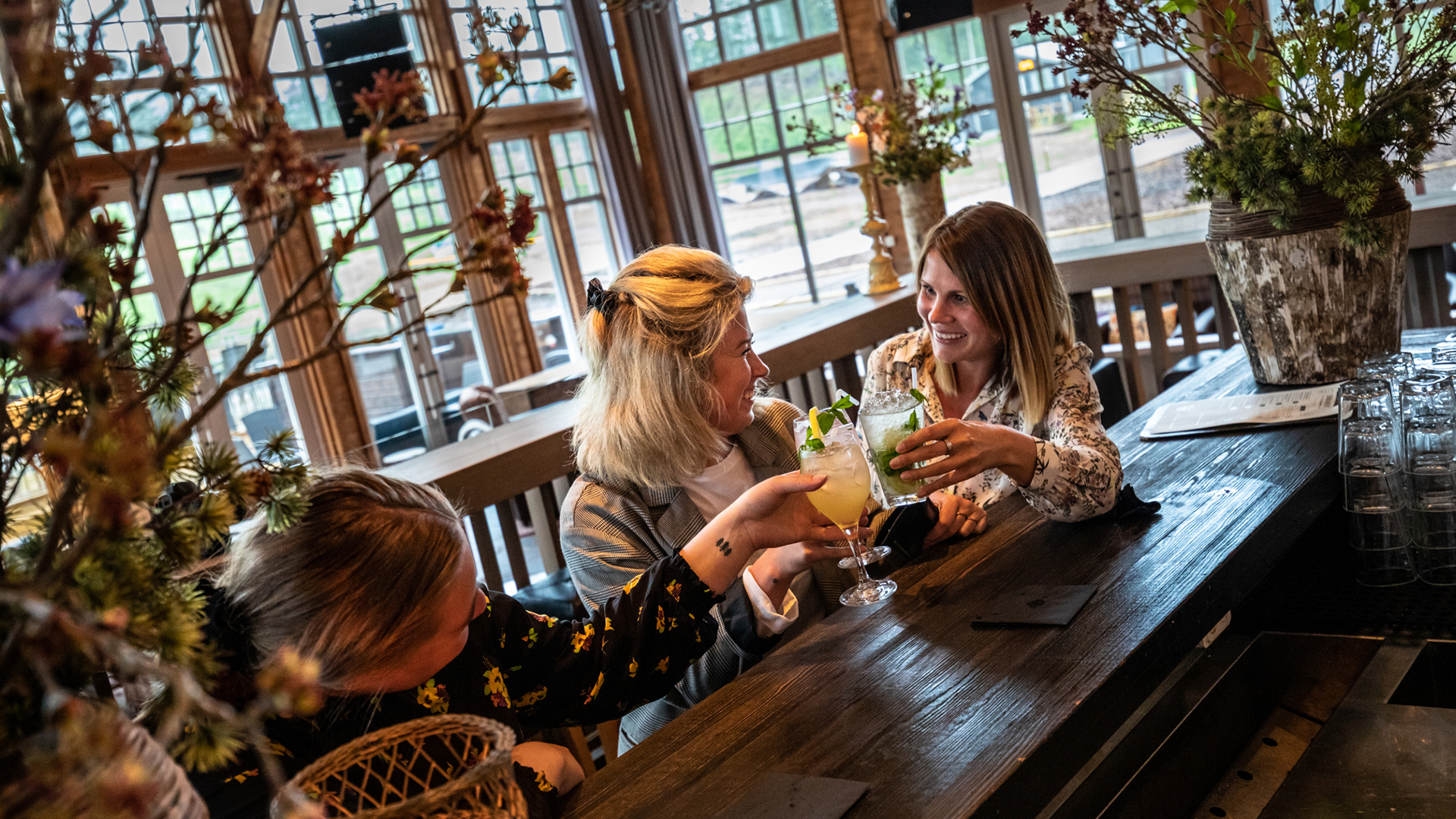 Tre kvinder skåler med drinks i en hyggelig bar, omgivet af blomster og grønne planter