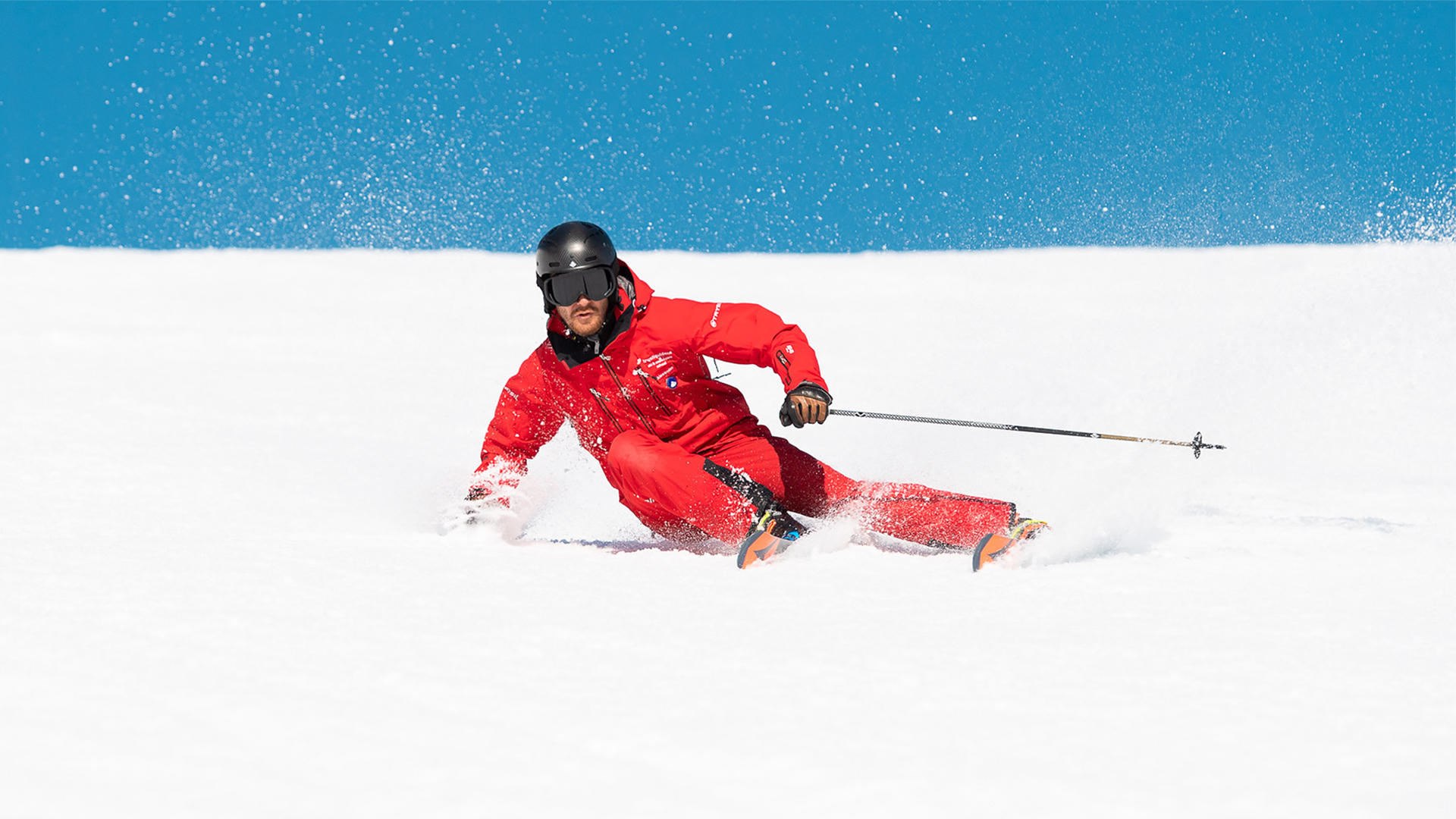 Man i röd skidklädsel åker skidor snabbt nerför en snötäckt backe under klarblå himmel