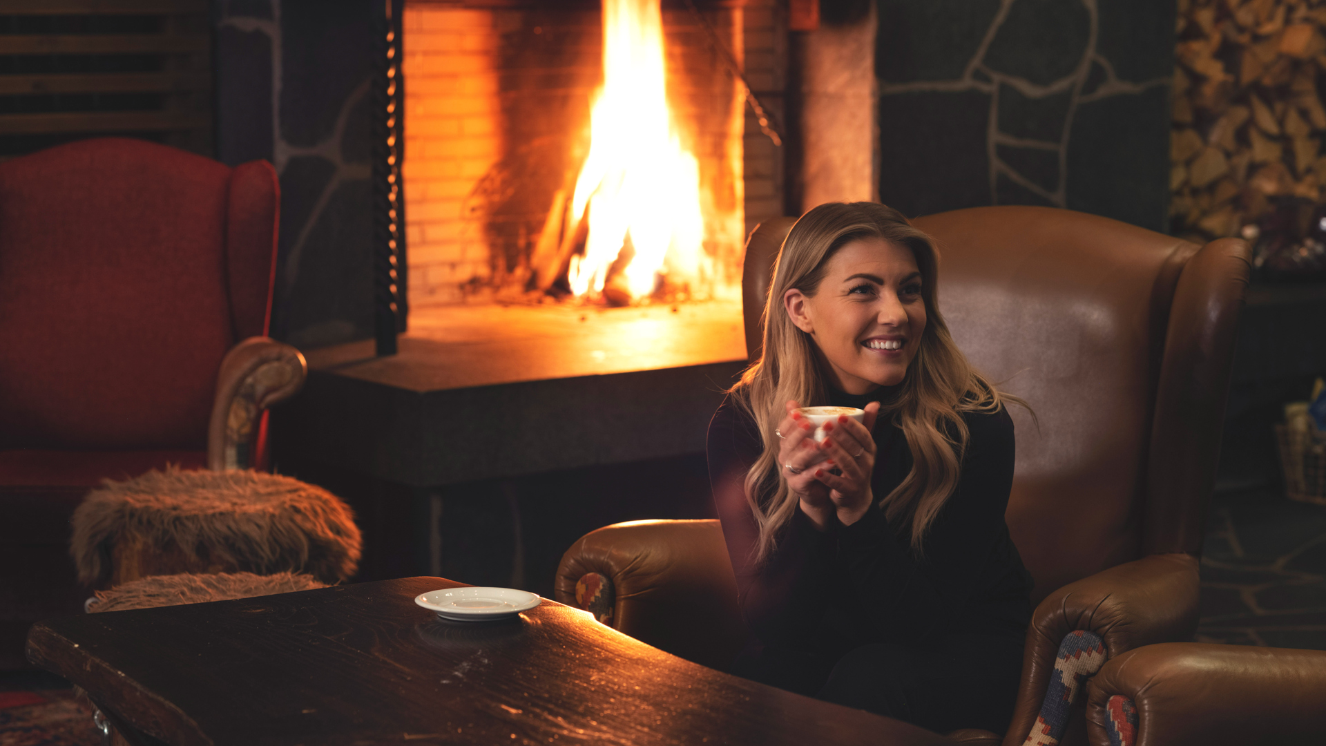 Kvinna som ler och håller i en kopp te framför en öppen spis i ett mysigt vardagsrum med brinnande eld