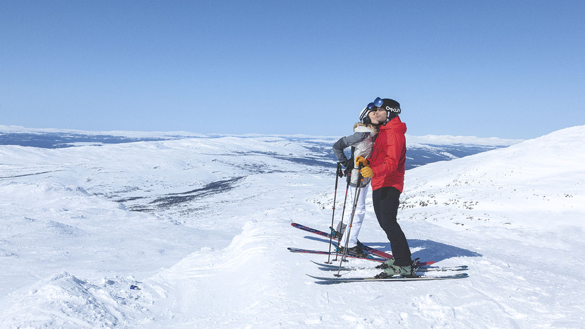 Par som åker skidor i fjällen, omgivna av snötäckta berg och klarblå himmel