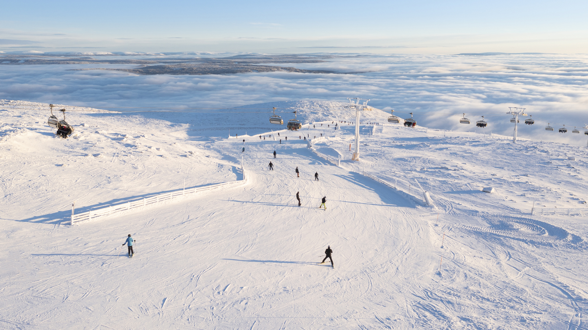 Skidåkare på fjälltoppen i Trysil, Norge, med snöklädda backar och bergstoppar under en klar himmel. Skidliftar syns i bakgrunden