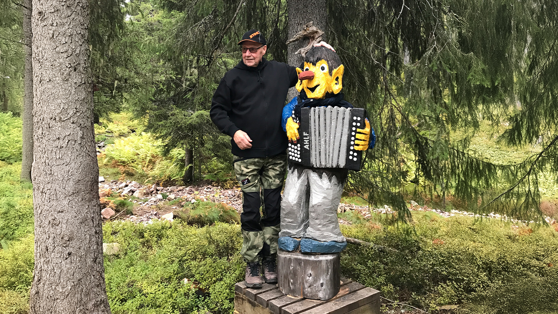 Man står bredvid en träskulptur av en figur som spelar dragspel i en skog