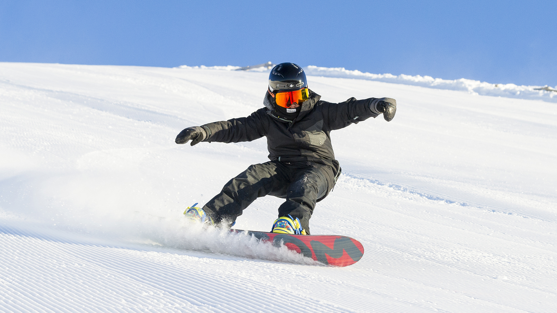 Snowboardåkare i svart dräkt och hjälm, utförande en kurva på en solig snötäckt backe