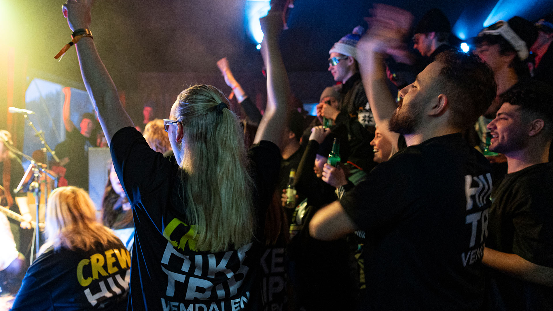 Folk nyder en koncert i en livlig indendørs atmosfære, iført sorte T-shirts. Stemningen er festlig med løftede hænder