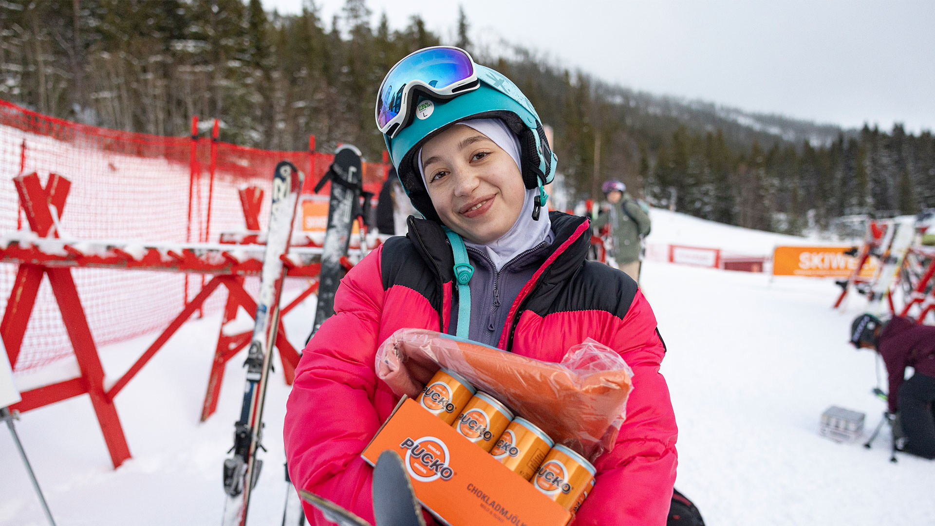 Skiløber i farverig jakke holder et seks-pack Pucko chokoladedrik og pølse på skisportssted, med snedækkede bjerge og ski i baggrunden