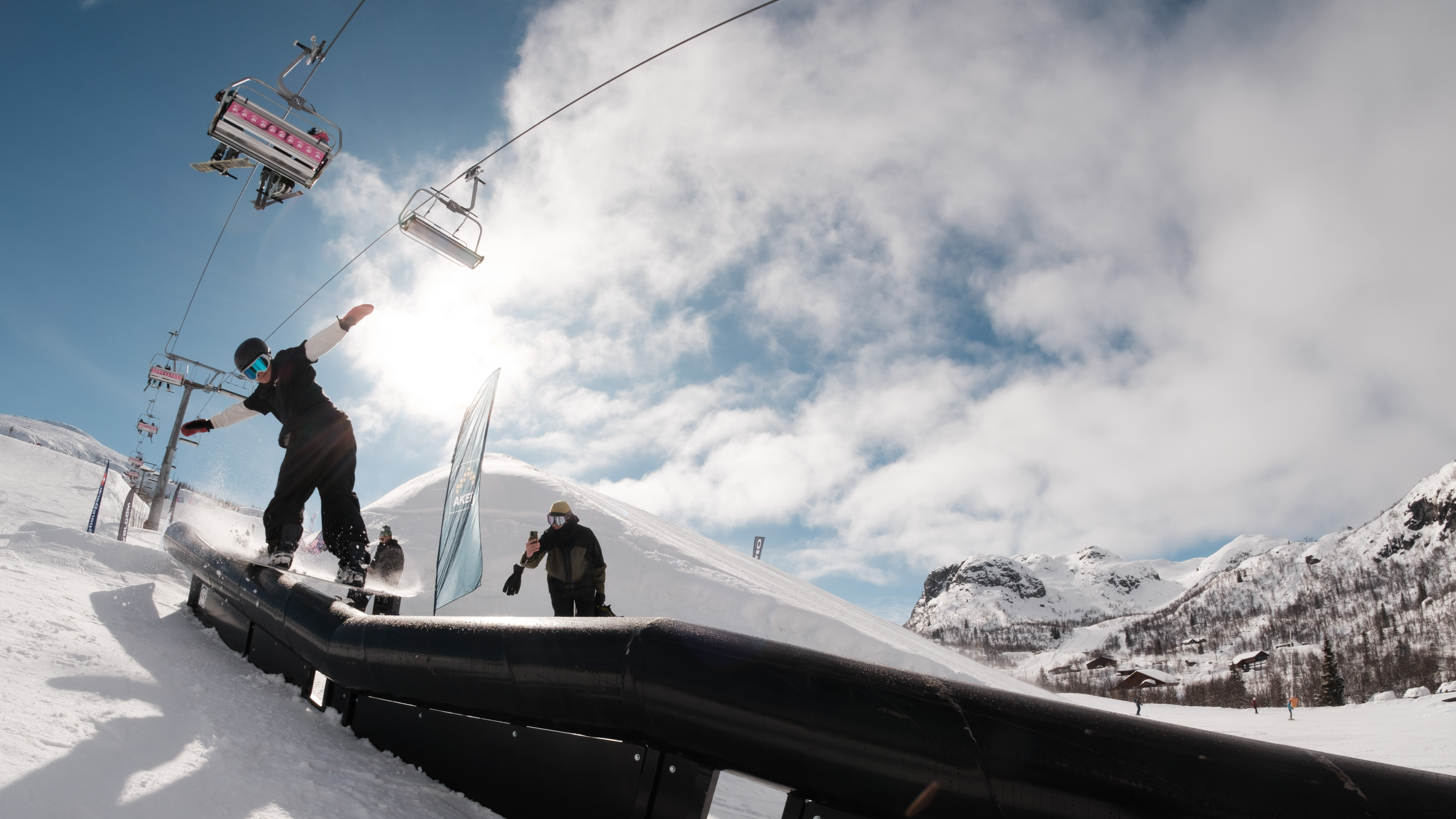 Snowboardåkare utför trick på rail i skidort, med sittlift och snötäckta berg i bakgrunden under en solig vinterdag