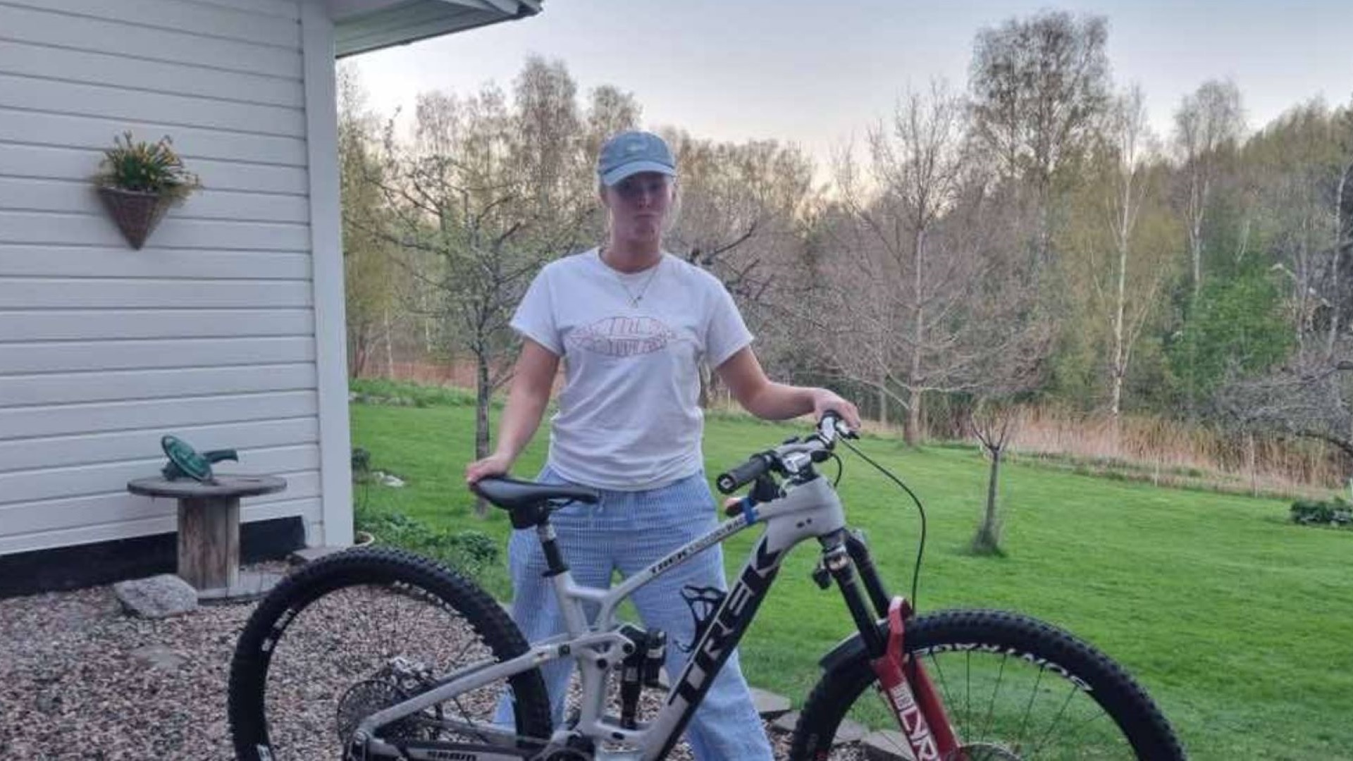 Kvinde iført afslappet tøj står ved siden af en Trek-cykel i en grøn have, omgivet af træer og en hvid bygning