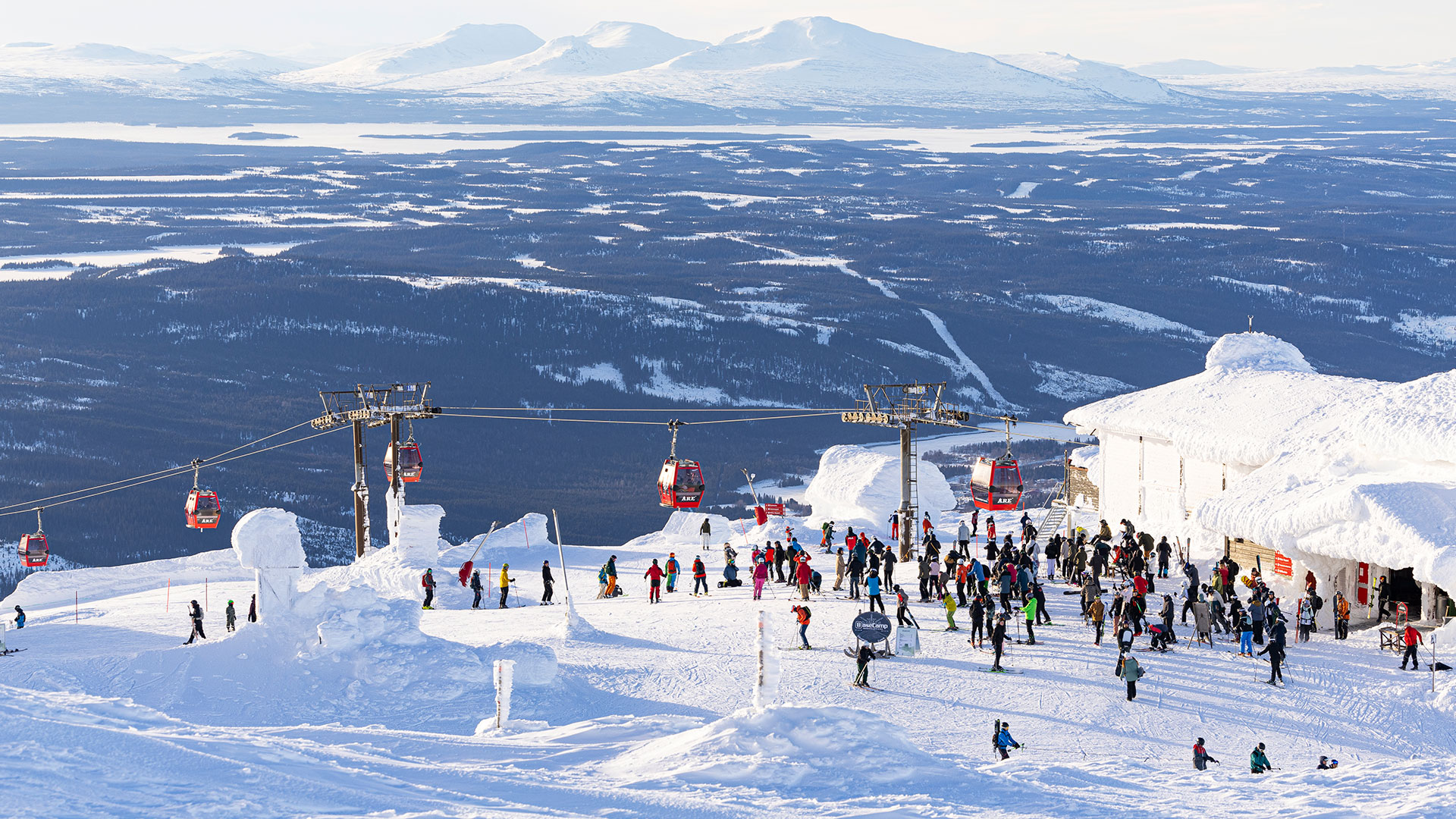 Skiers gather at a snow-covered cabin at the top of Åreskutan, with a mountain landscape in the background and a cable car in operation. A perfect winter destination for skiing in Sweden