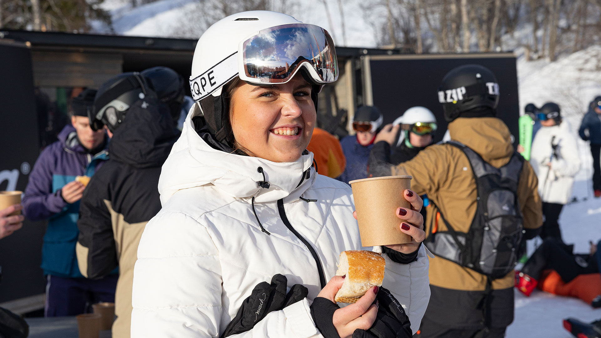 A woman in ski clothing is holding a coffee mug and a pastry at a winter resort