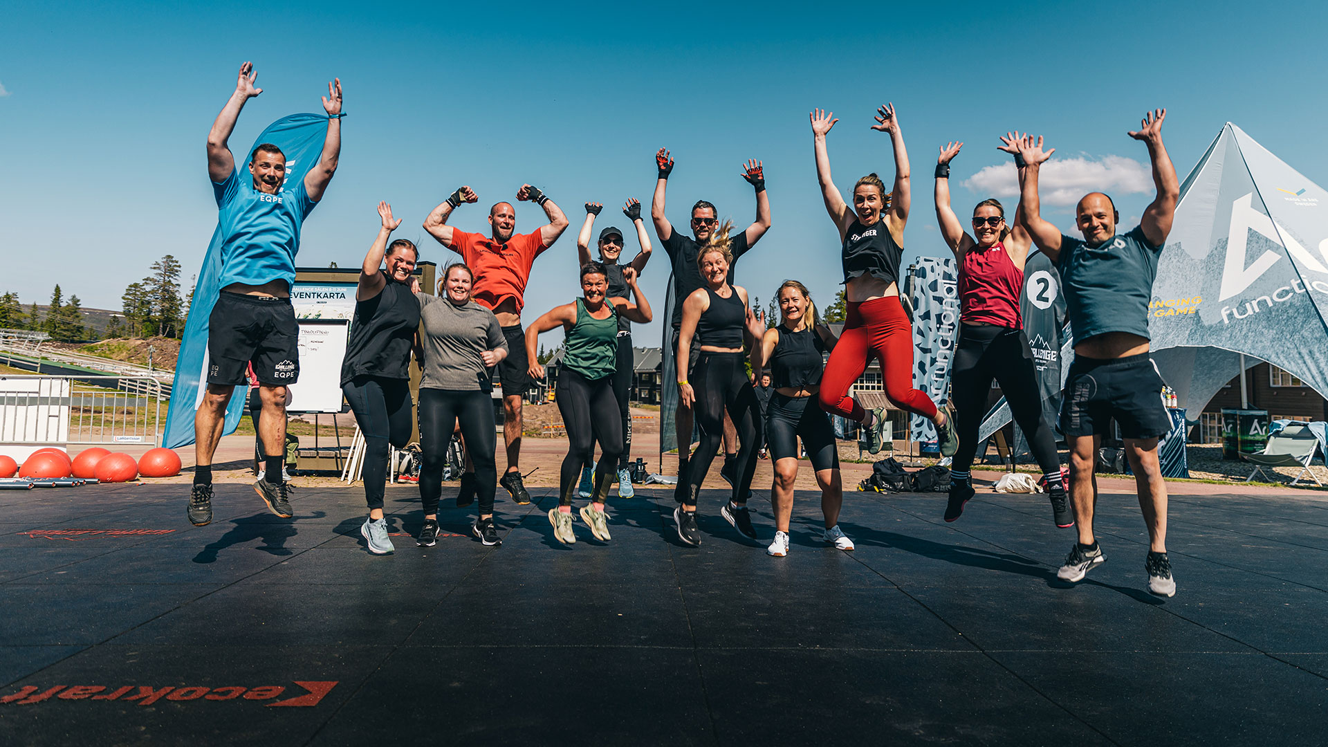 Gruppetreningsøkt utendørs med glade deltakere som hopper på en solfylt dag, omgitt av treningsutstyr og flagg