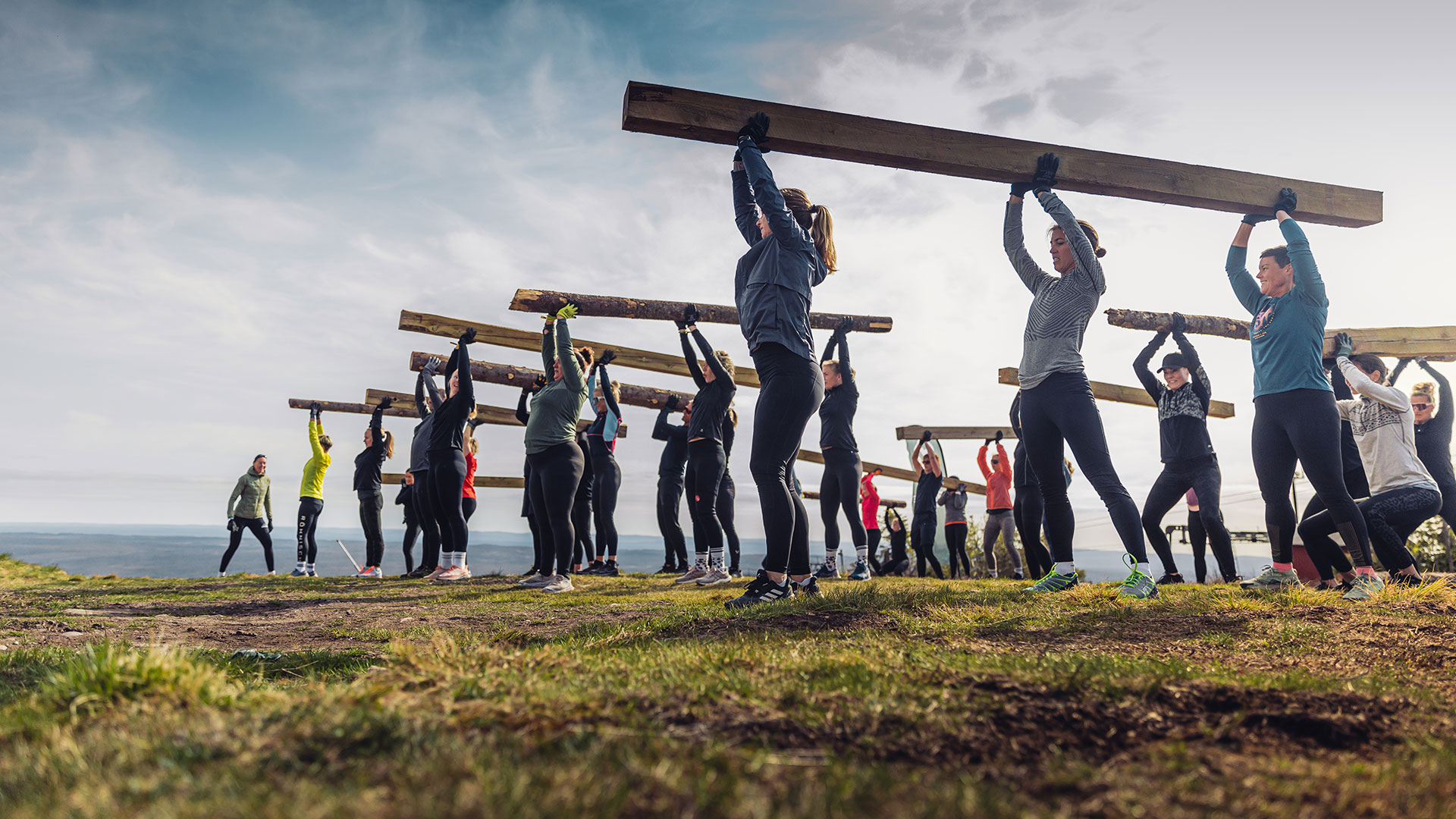 Personer deltar i gruppetrening utendørs og løfter trebjelker over hodet på en grønn eng under en skyet himmel
