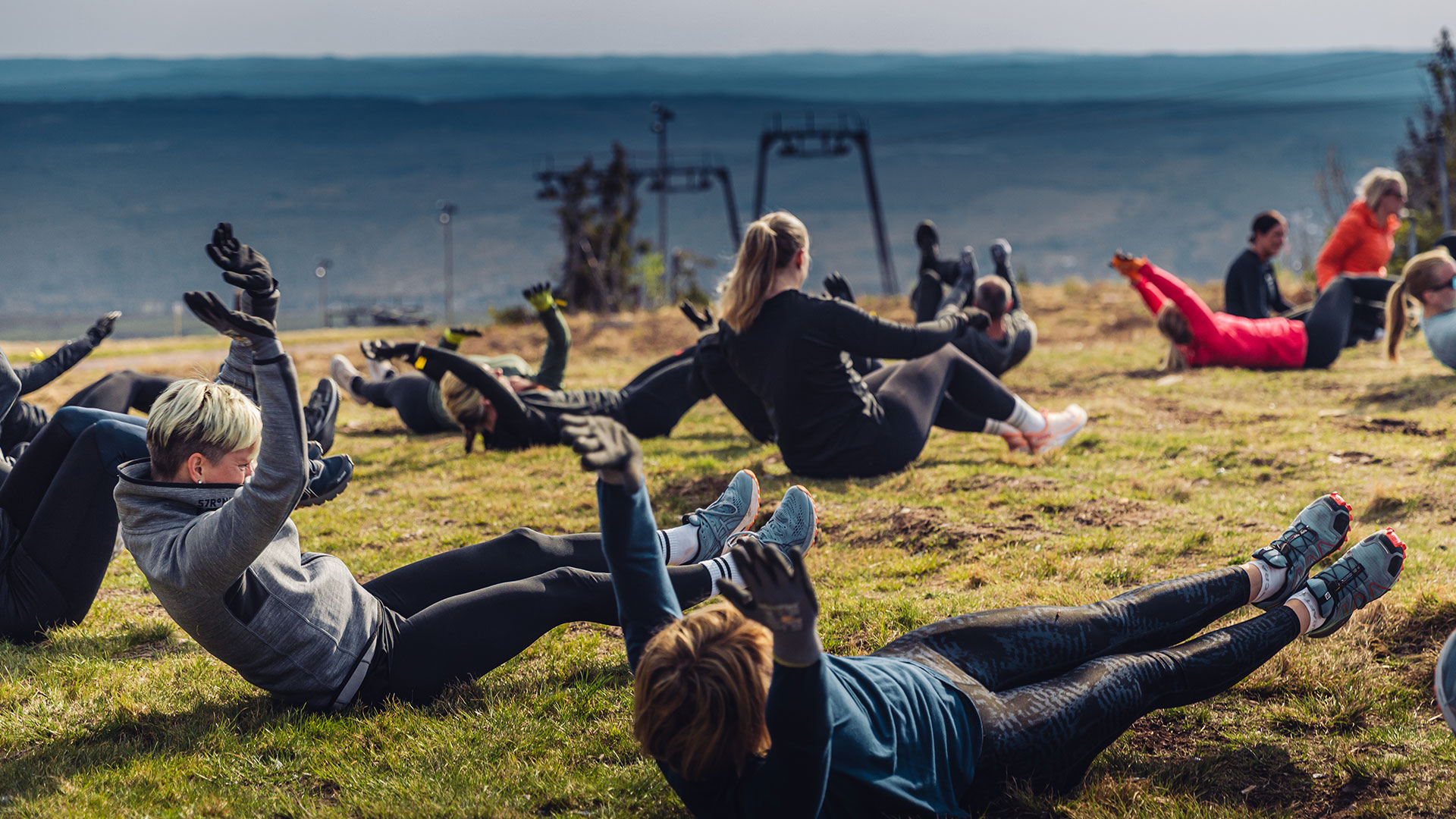 Mennesker deltar i gruppetrening utendørs på en gresskledd skråning med utsikt over landskapet