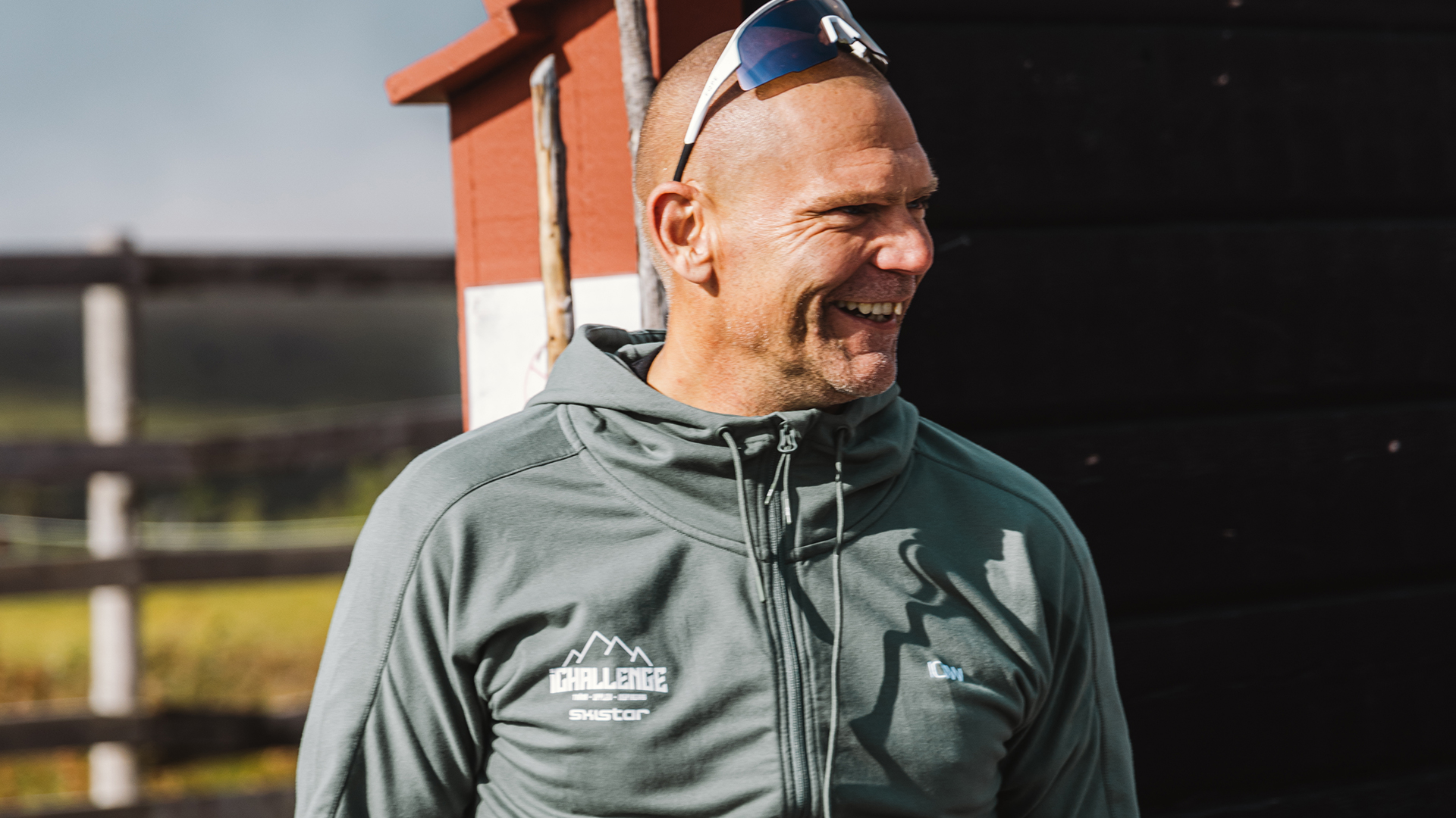 Man i grå jacka med "Challenge" logotyp, står leende utomhus i soligt väder