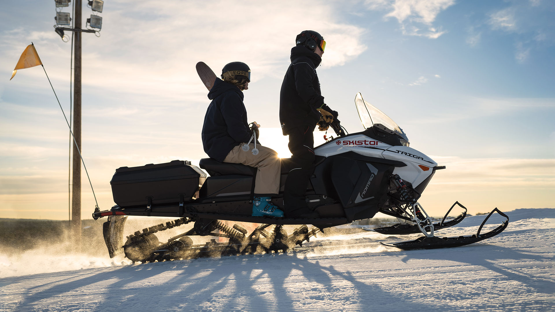Två personer åker snöskoter på ett snötäckt fält under en klar himmel, med en flagga på bakdelen av skotern