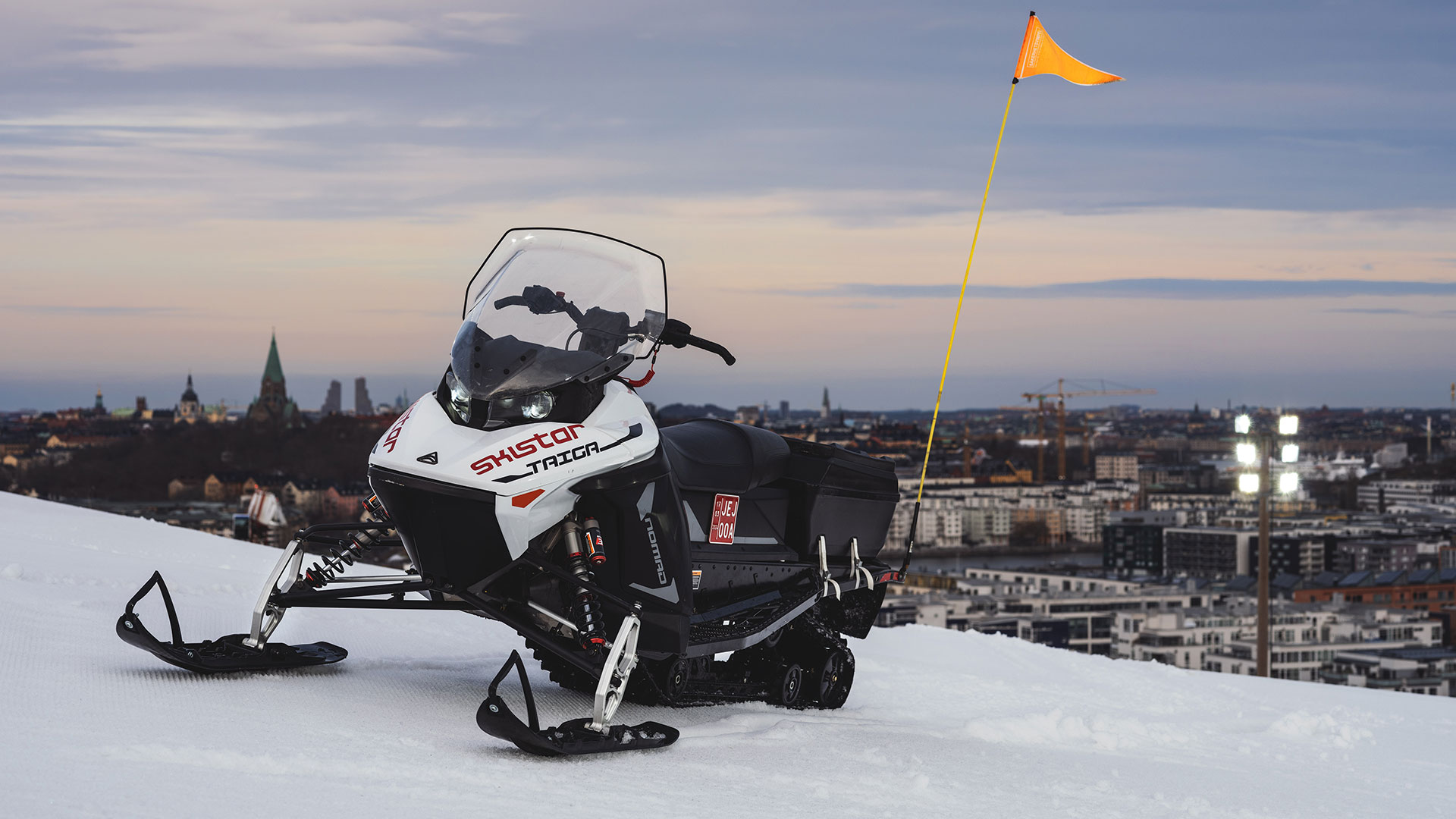 Snöskoter med varningsflagga på en snötäckt backe över en stadssilhuett i skymningen