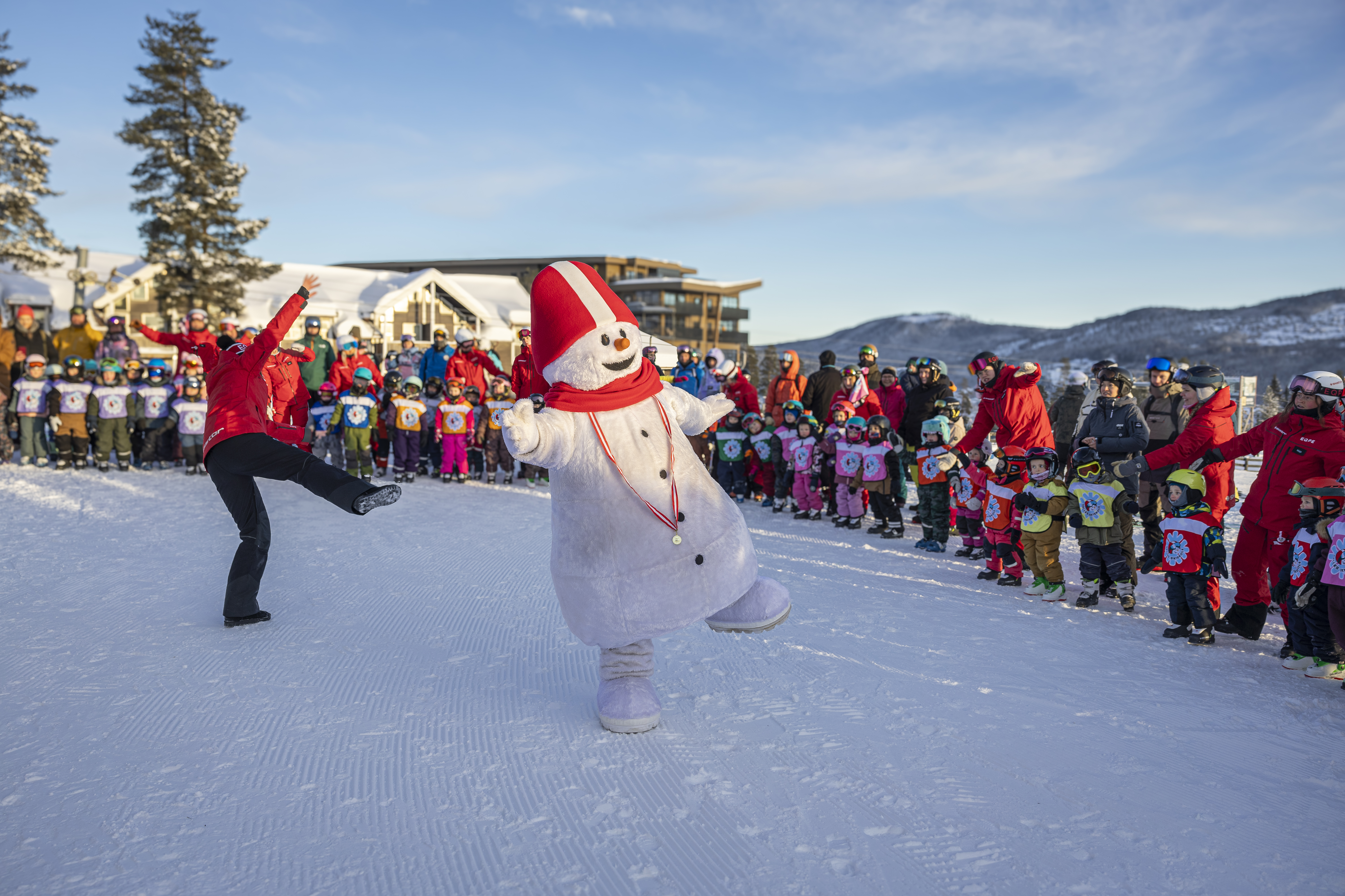 Snögubbe och skidlärare dansar framför en grupp barn på en skidort, omgivna av vintrigt landskap och personal i röda jackor