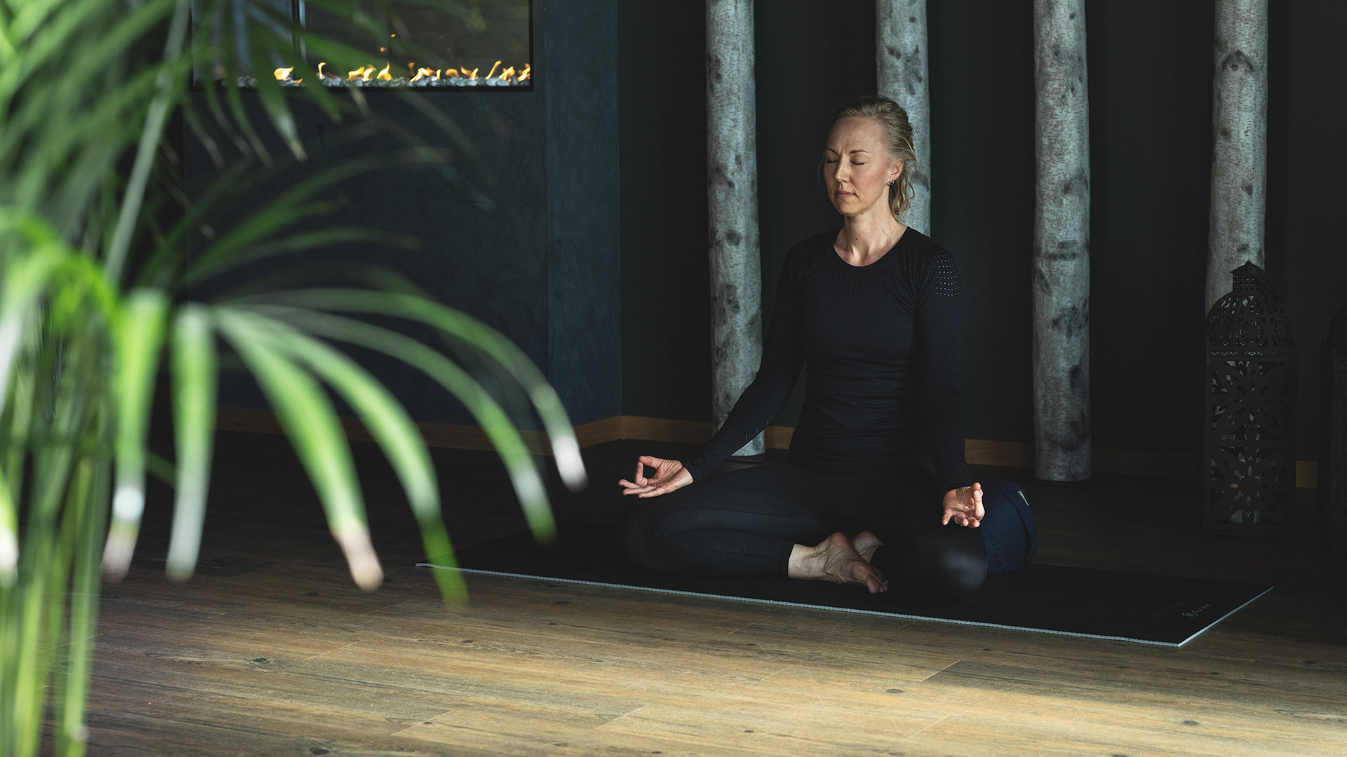 Kvinna mediterar på en yogamatta i ett lugnt rum, omgiven av naturliga element som växter och trädstammar