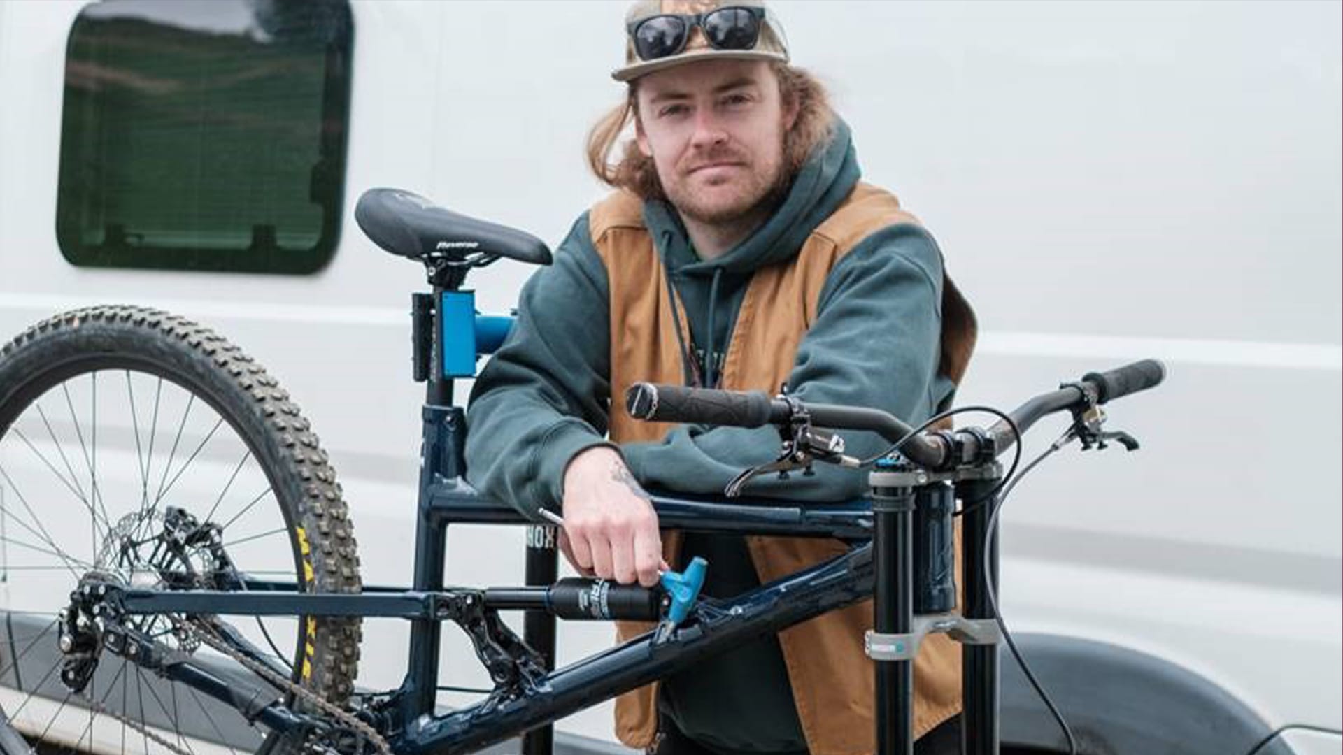 Mand står med mountainbike. Han bærer solbriller, kasket, hættetrøje og vest