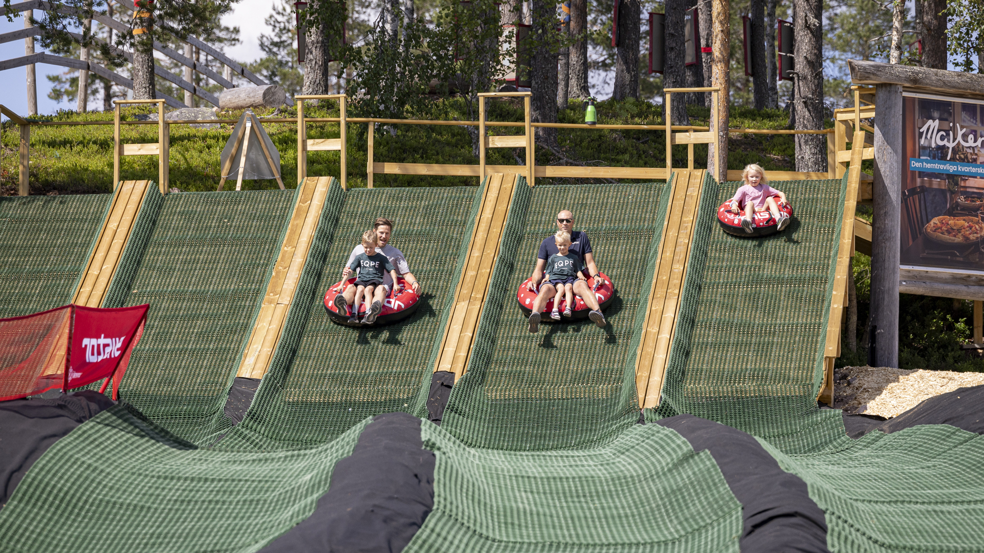 Personer åker nerför en grön konstgrässbacke på uppblåsbara tuber i en äventyrspark omgiven av skog