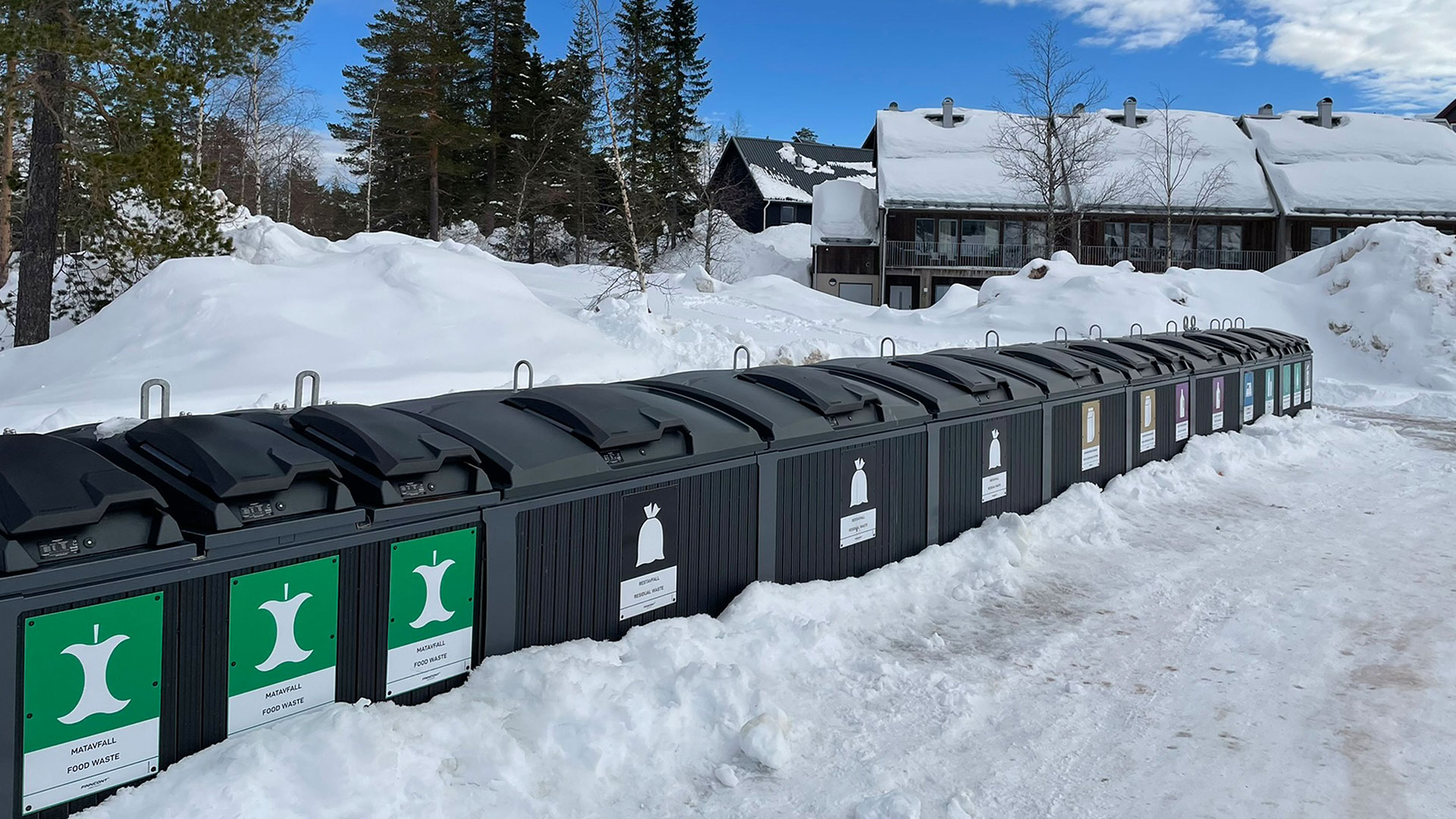 Rader med återvinningskärl för matavfall och andra material i ett vintrigt landskap vid en byggnad, omgivet av snö och träd