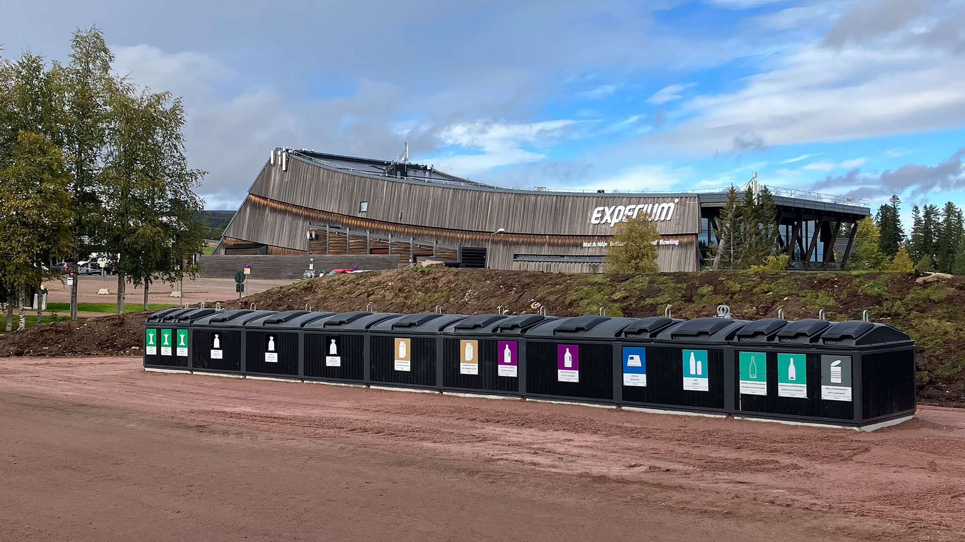 Återvinningsstation med moderna containrar framför en byggnad med skylt "Experium" och träd i bakgrunden