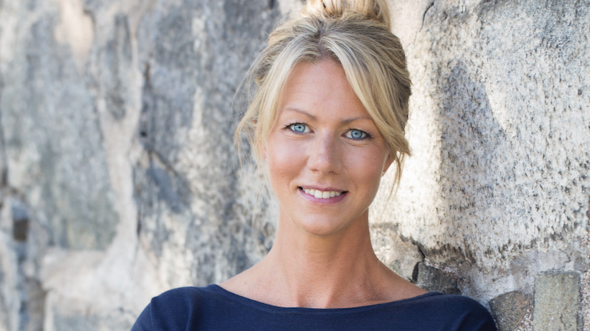 Kvinna med blont hår och blå klänning står mot en stenvägg, ler mot kameran