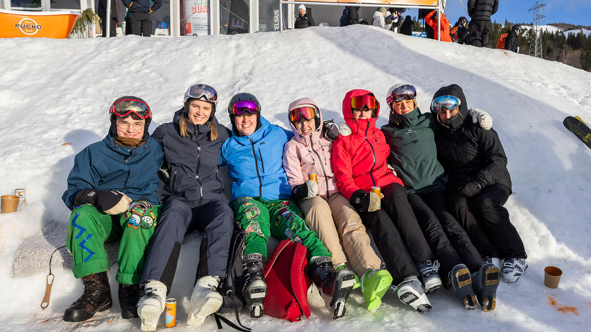 Grupp av glada människor i skidkläder sitter på snön vid en skidbacke