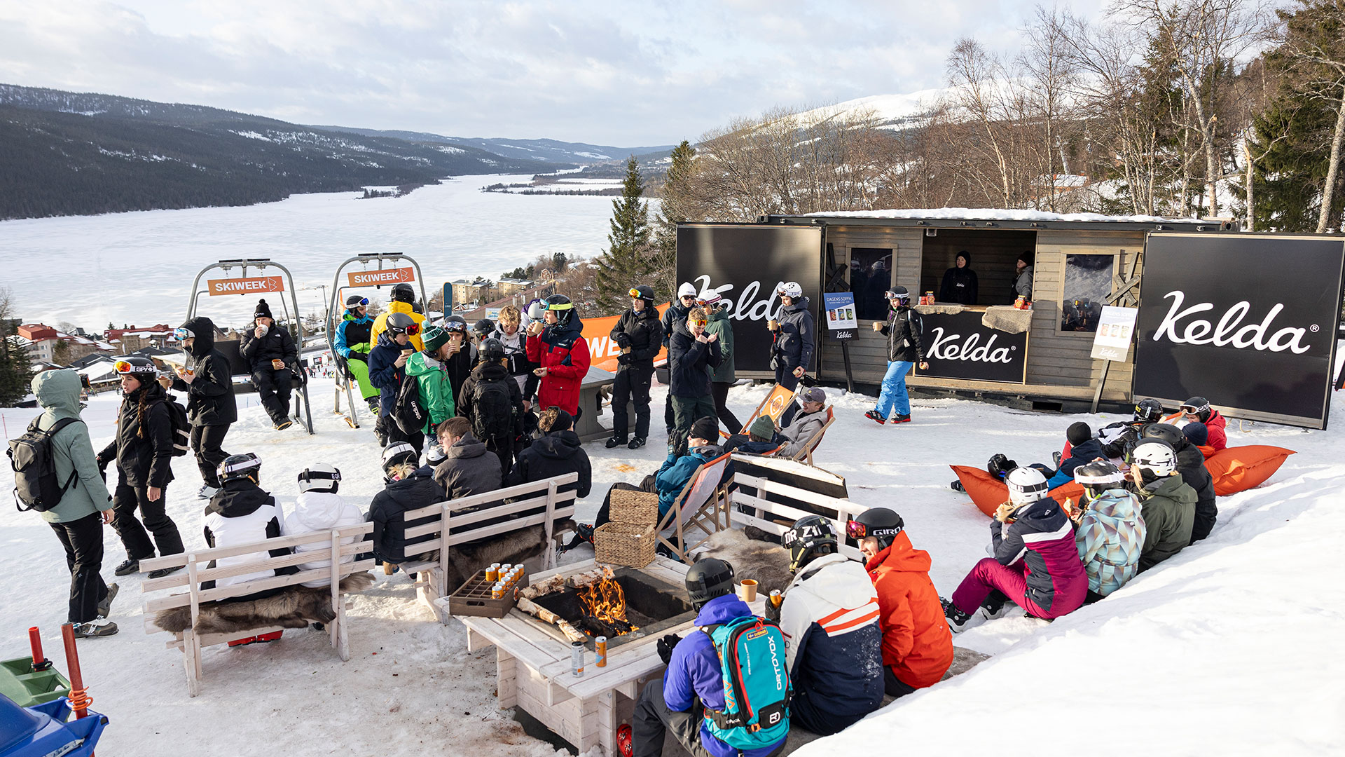 Personer njuter av vinteraktiviteter vid en skidanläggning i Åre, Sverige. Det finns sittplatser nära en eld och skyltar med varumärket Kelda syns i bakgrunden. En vacker snötäckt landskap och fryst sjö sträcker sig i fjärran