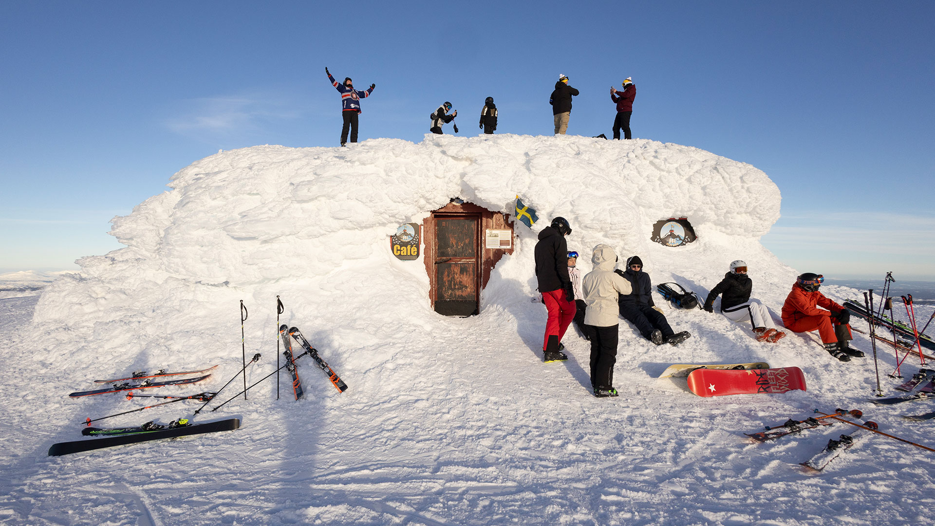 Personer samlas vid ett snötäckt café på en bergstopp, med skidor och snowboard runt omkring. Klarblå himmel i bakgrunden