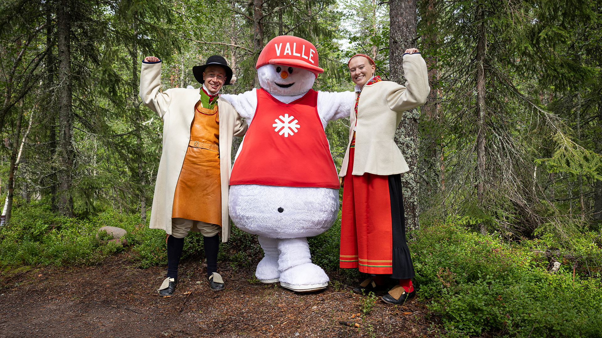 Maskot ved navn Valle står i midten af en skov mellem to personer i traditionelt tøj. De smiler og løfter armene i en munter gestus