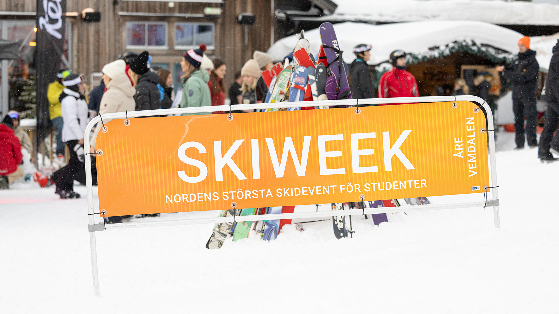 Skylt för Skiweek i Åre och Vemdalen, Nordens största skidevent för studenter, med människor och skidutrustning i bakgrunden