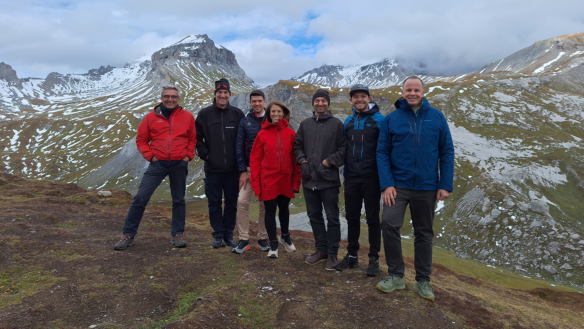Grupp personer står i alperna, klädda i varma kläder, med snöbeklädda berg i bakgrunden under en molnig himmel