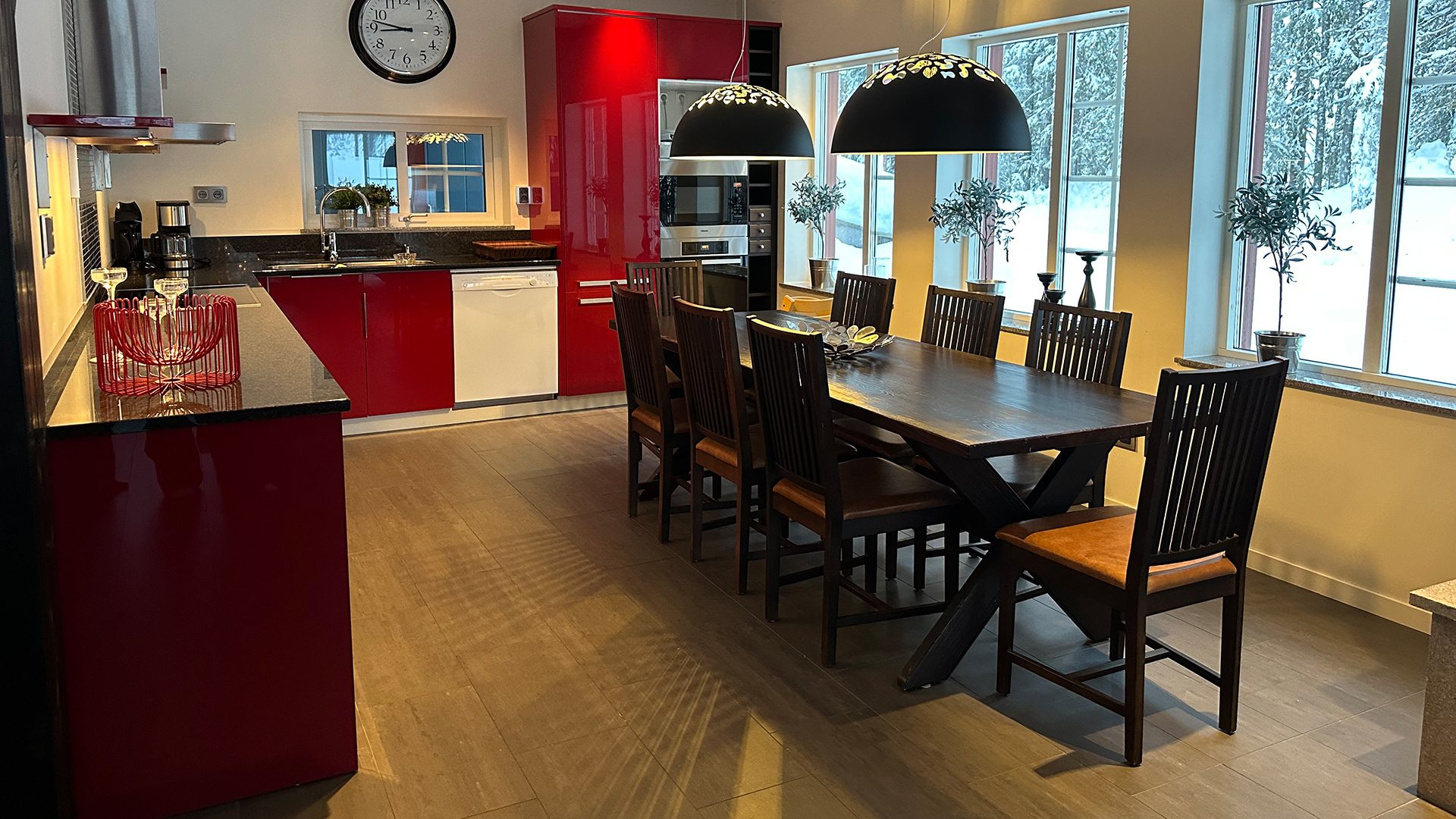 Modernt kök med röda skåp och träbord, omgiven av stolar. Svarta bänkskivor och stora fönster som släpper in naturligt ljus. Hängande lampor över matbordet