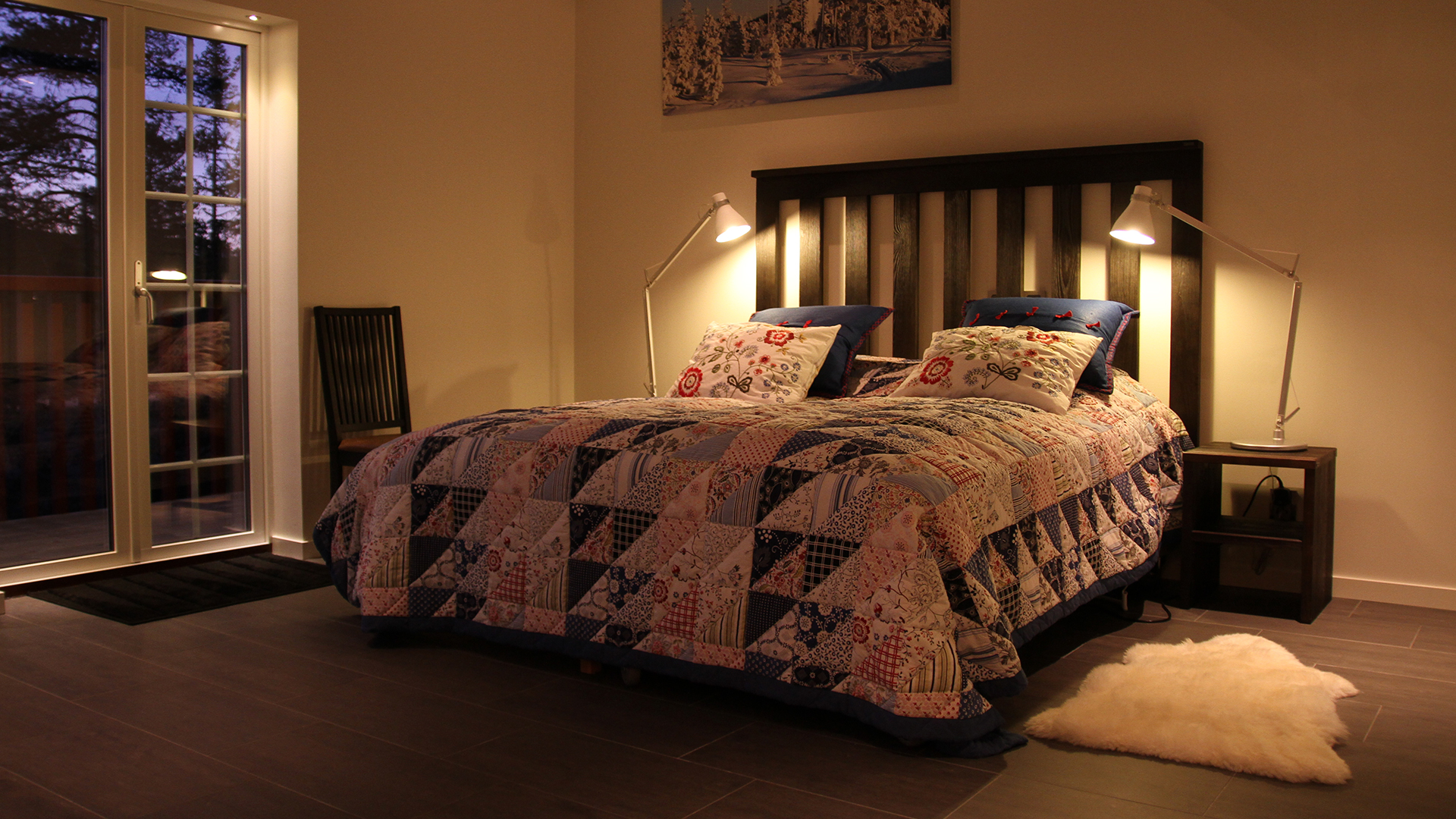 Sovrum med dubbelsäng, täckt av ett mönstrat täcke, två sänglampor och en matta på golvet. Fransk dörr med utsikt mot kvällshimmel