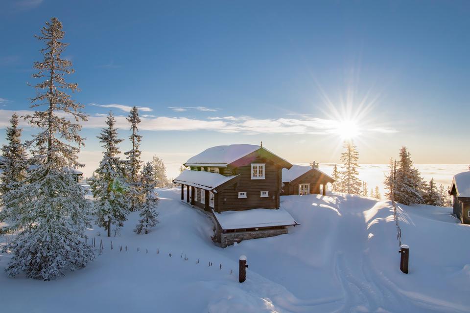 Træhytte i sneklædt landskab med solnedgang, omgivet af snedækkede træer og blå himmel, skaber en stemningsfuld vinteridyl