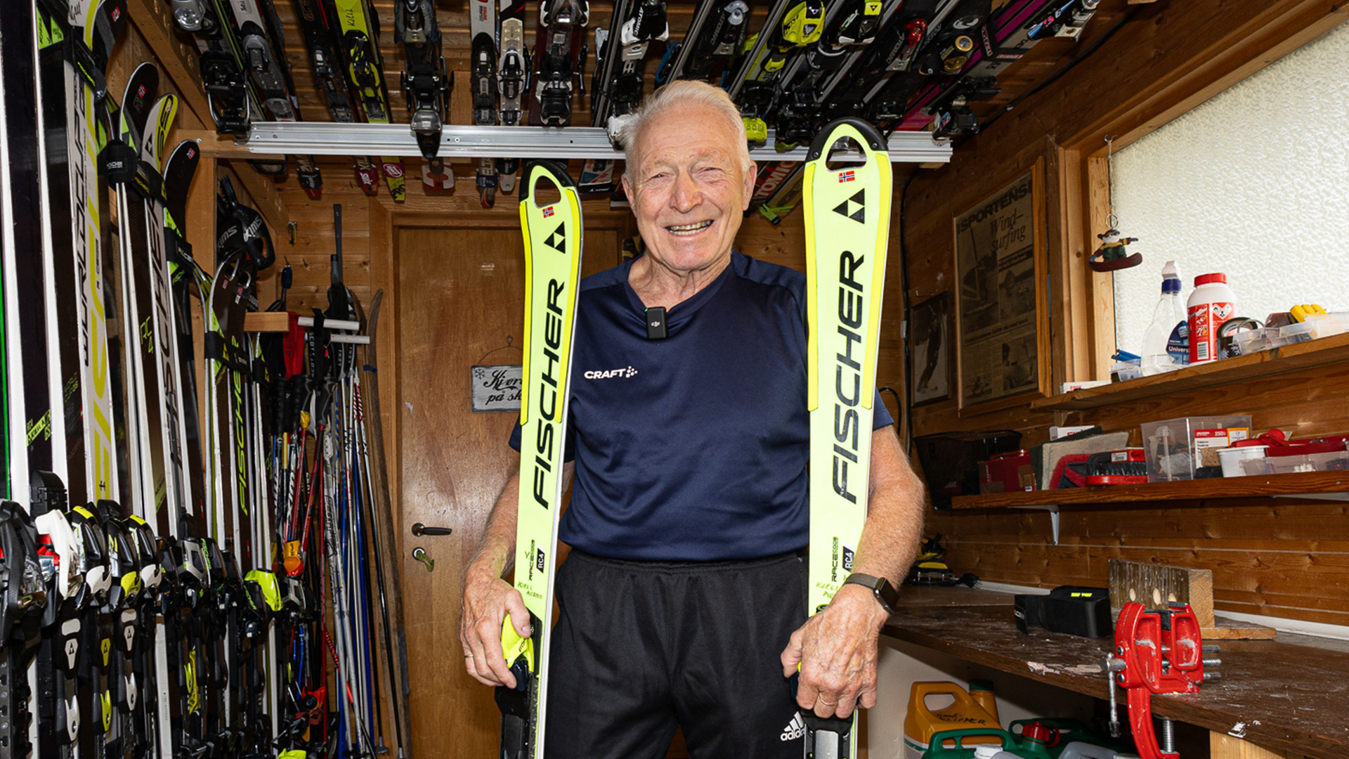 Ældre mand i skirum omgivet af ski, holder et par gule Fischer-ski