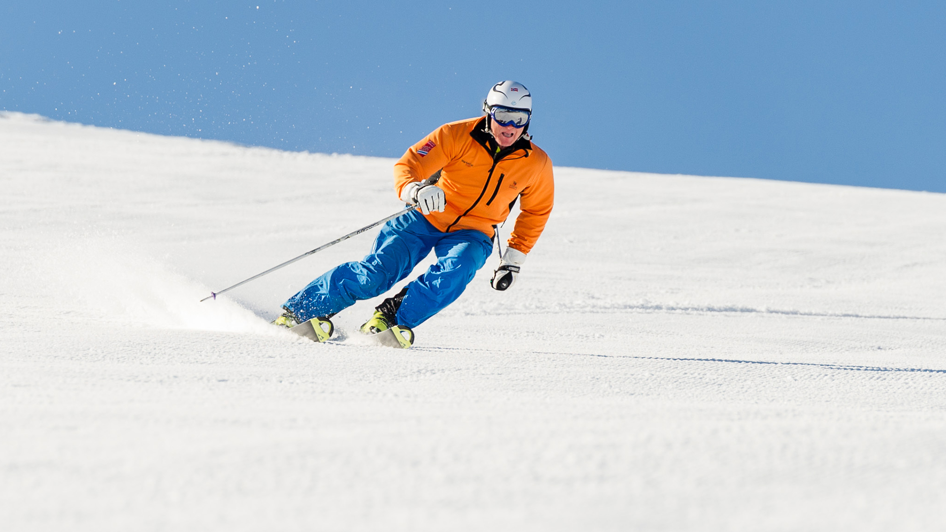 A person skiing down a snow-covered slope in sunny weather, wearing an orange jacket and blue pants
