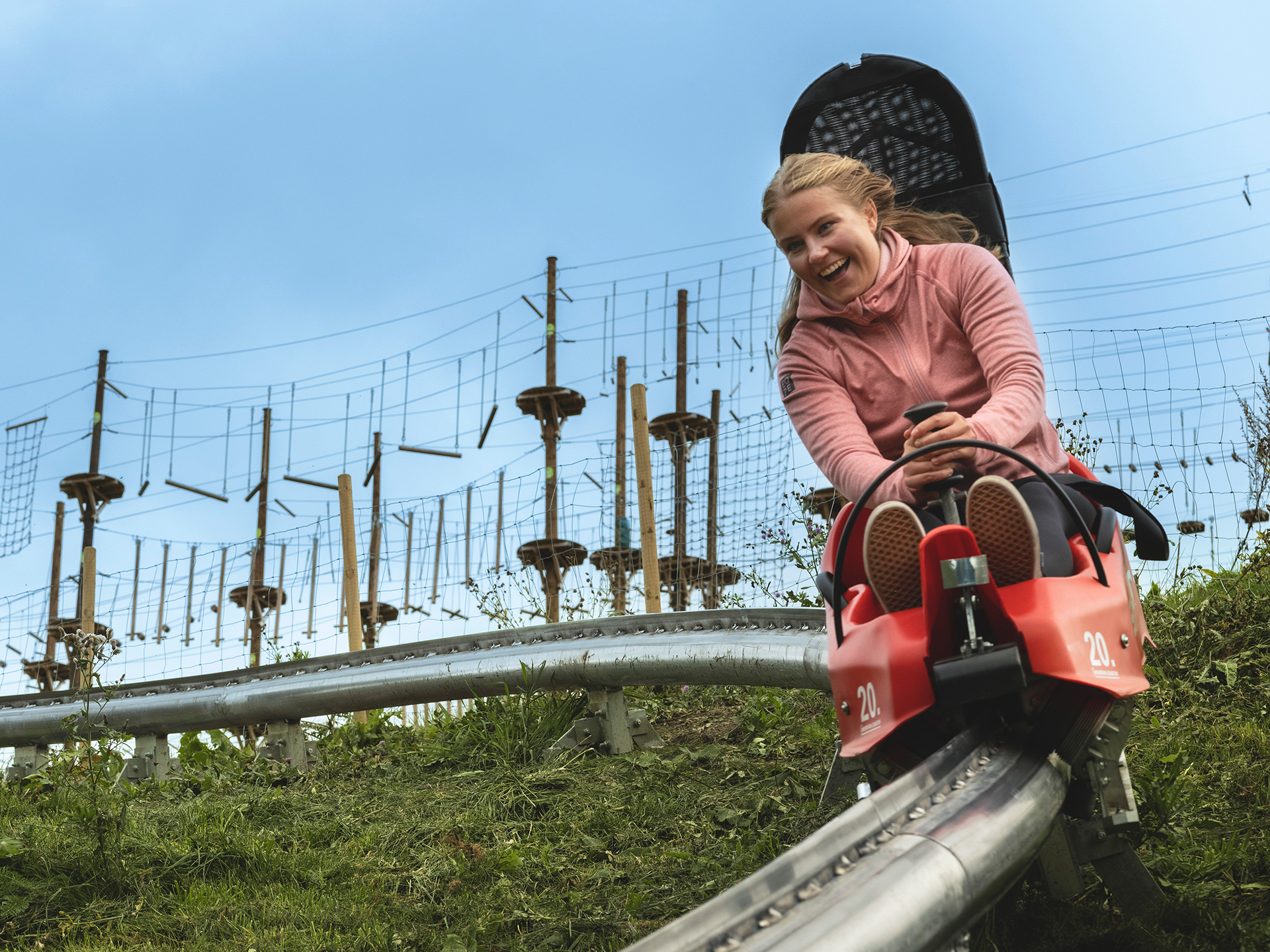 Kvinna åker rodel på en bergbana utomhus, med trädklättringsbanor i bakgrunden under en klarblå himmel
