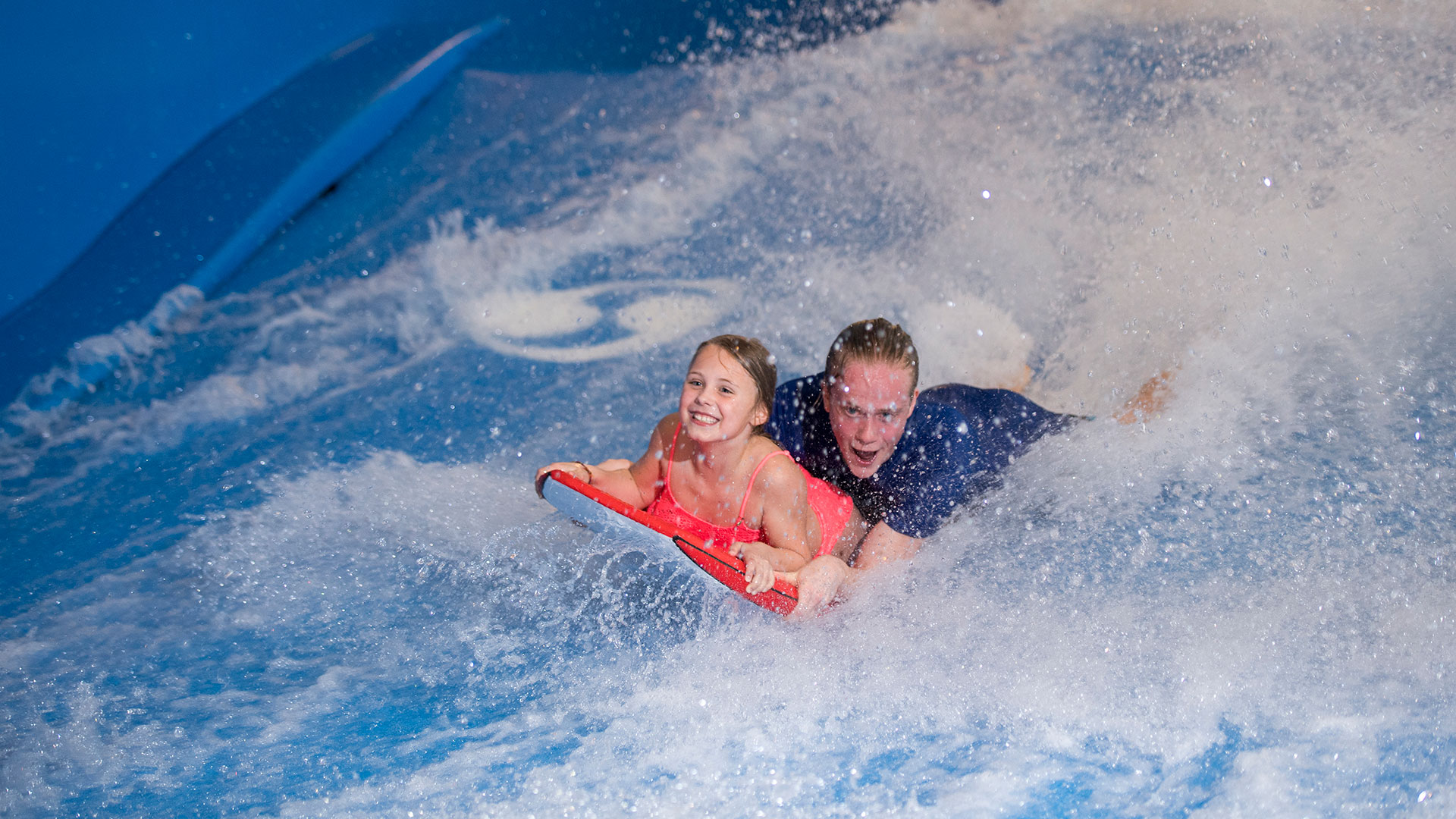 Barn og voksen kører på bodyboard på en bølge i en vandpark, ser glade ud og nyder aktiviteten