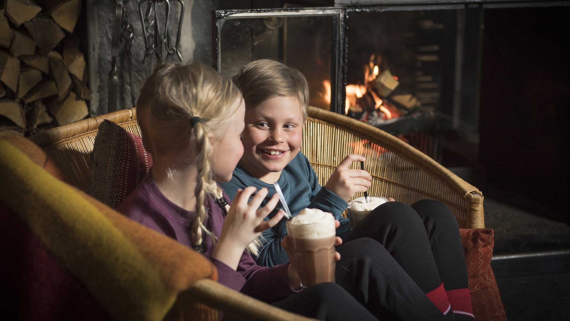 Två barn dricker varm choklad framför en sprakande brasa i en mysig korgsoffa